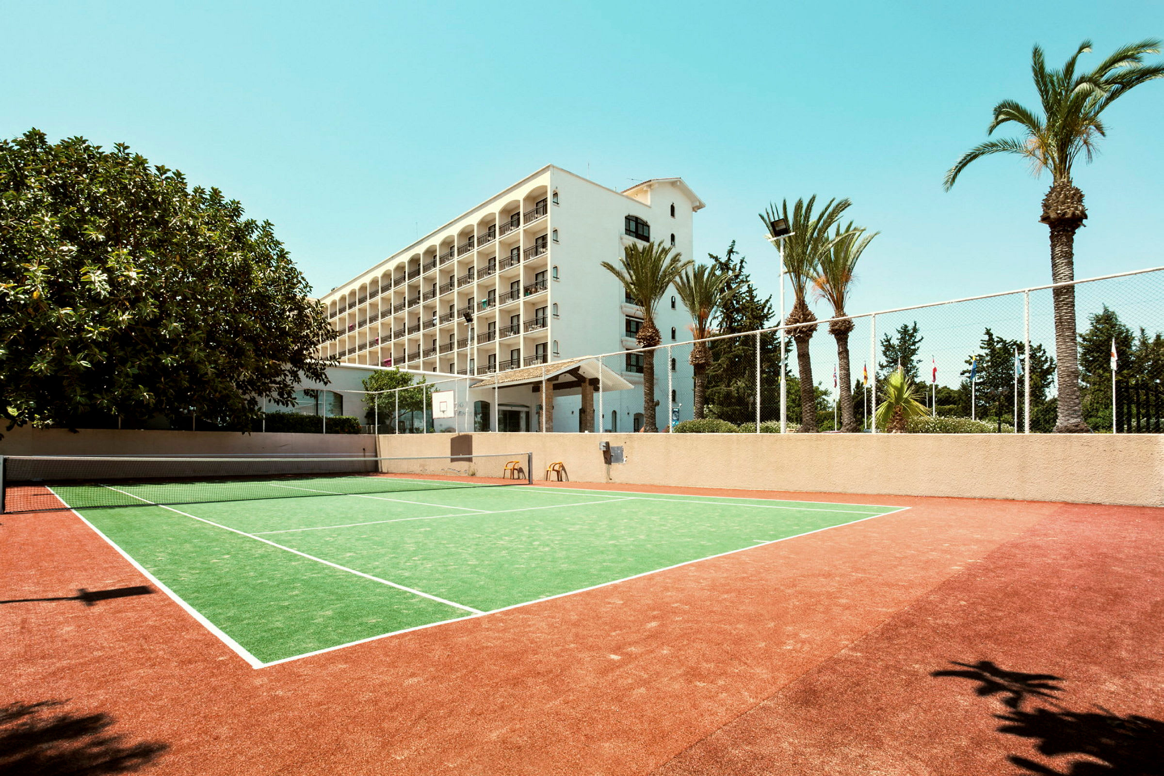 En tennisbane foran et hotell med flere etasjer og omgitt av palmer. (AI-generert tekst)