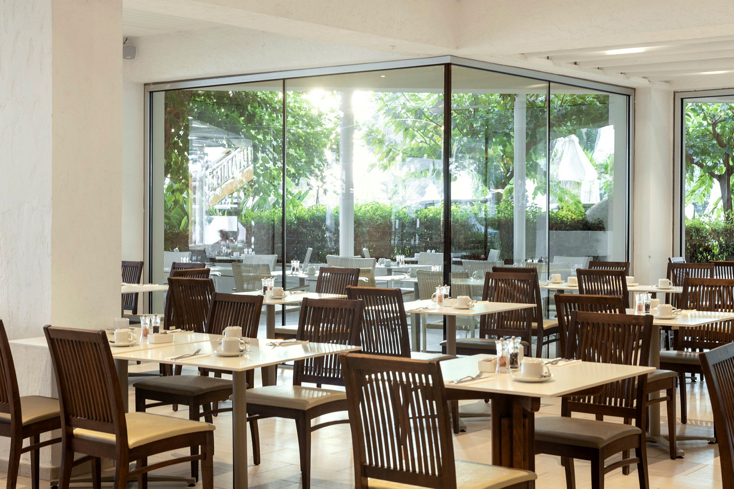 Bilde av en restaurant med trebord og stoler, samt utsikt mot grønne omgivelser gjennom store glassvinduer. (AI-generert tekst)