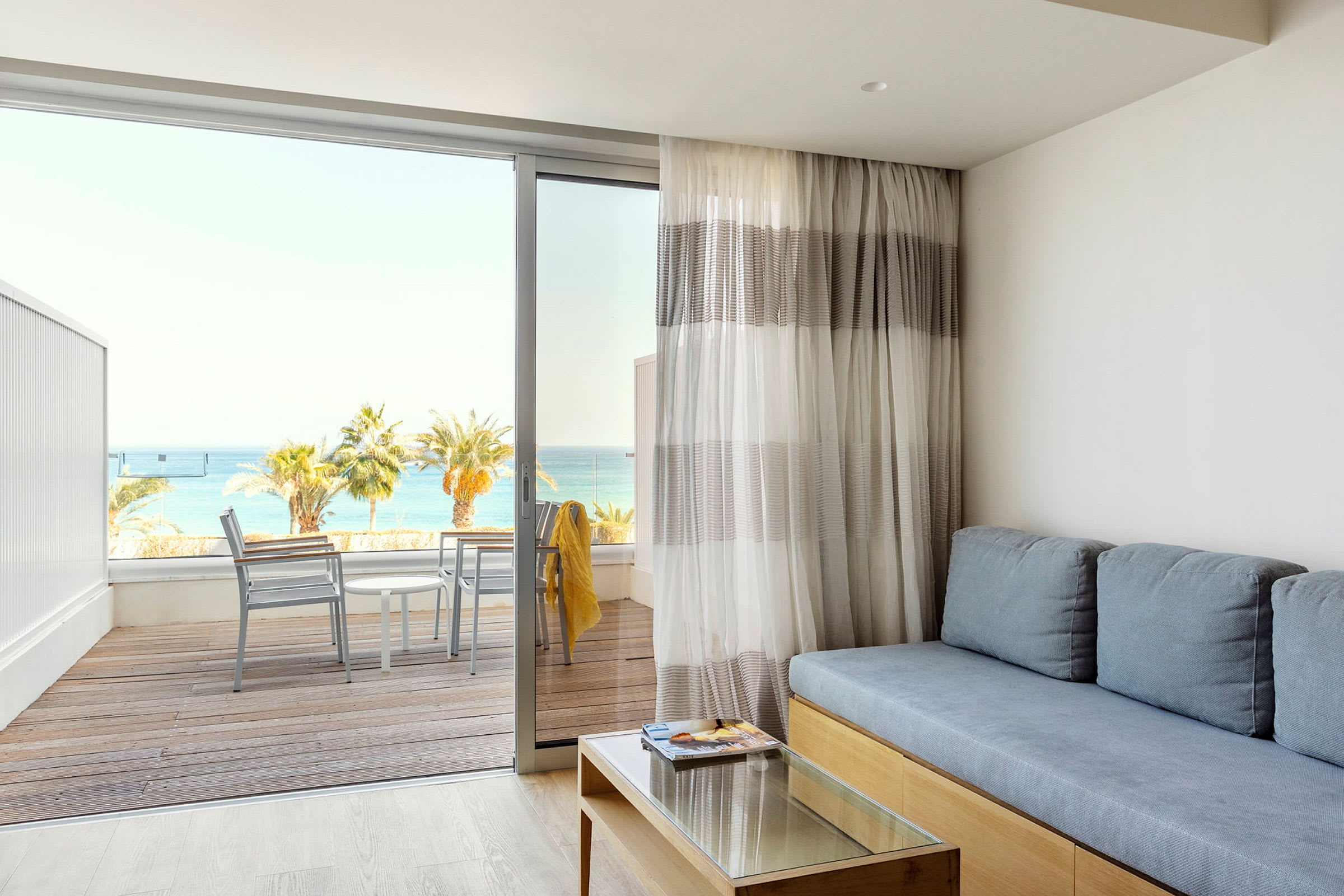 Bilde av en stue med en blå sofa, et glassbord og en balkong med utsikt over havet. (AI-generert tekst)