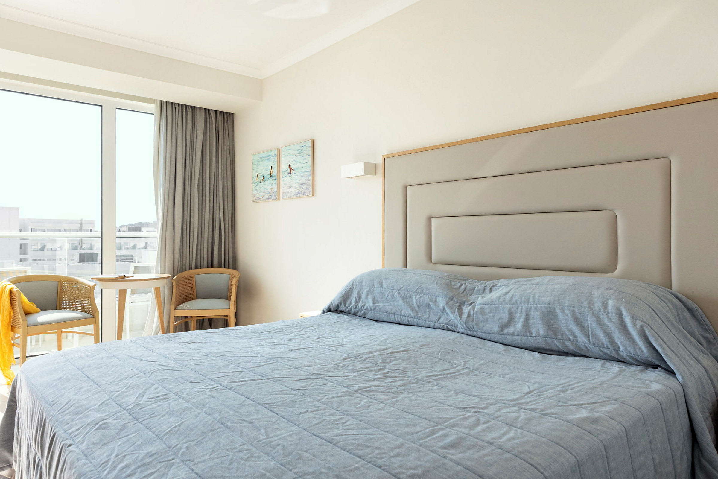 Et hotellrom med en seng redt med blått sengeteppe, to stoler og et bord nær et vindu. (AI-generert tekst)