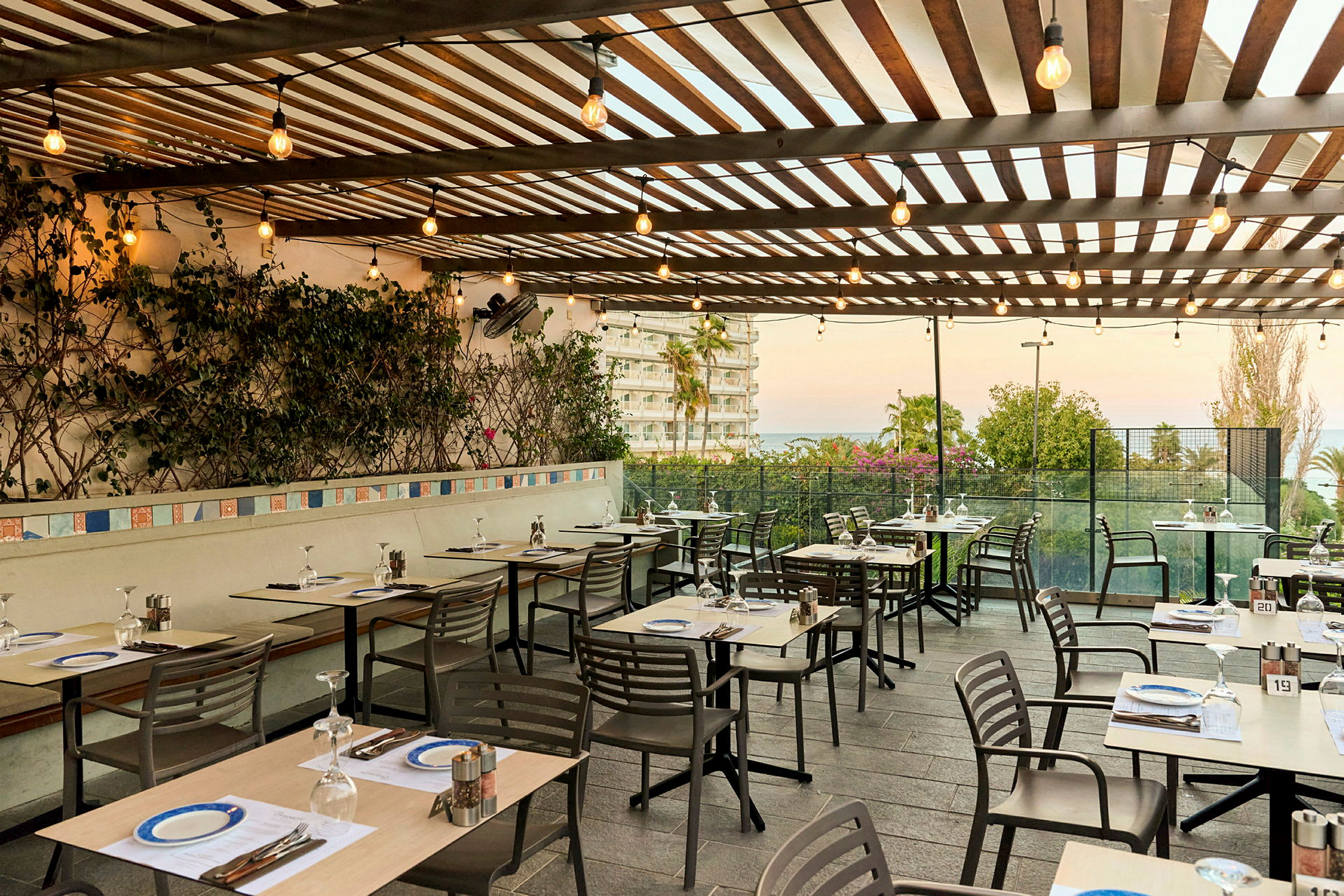 En utendørs restaurant med dekkede bord og stoler under en trepergola dekorert med planter og lyslenker. Utsikt over en bygning og hage mot havet. (AI-generert tekst)