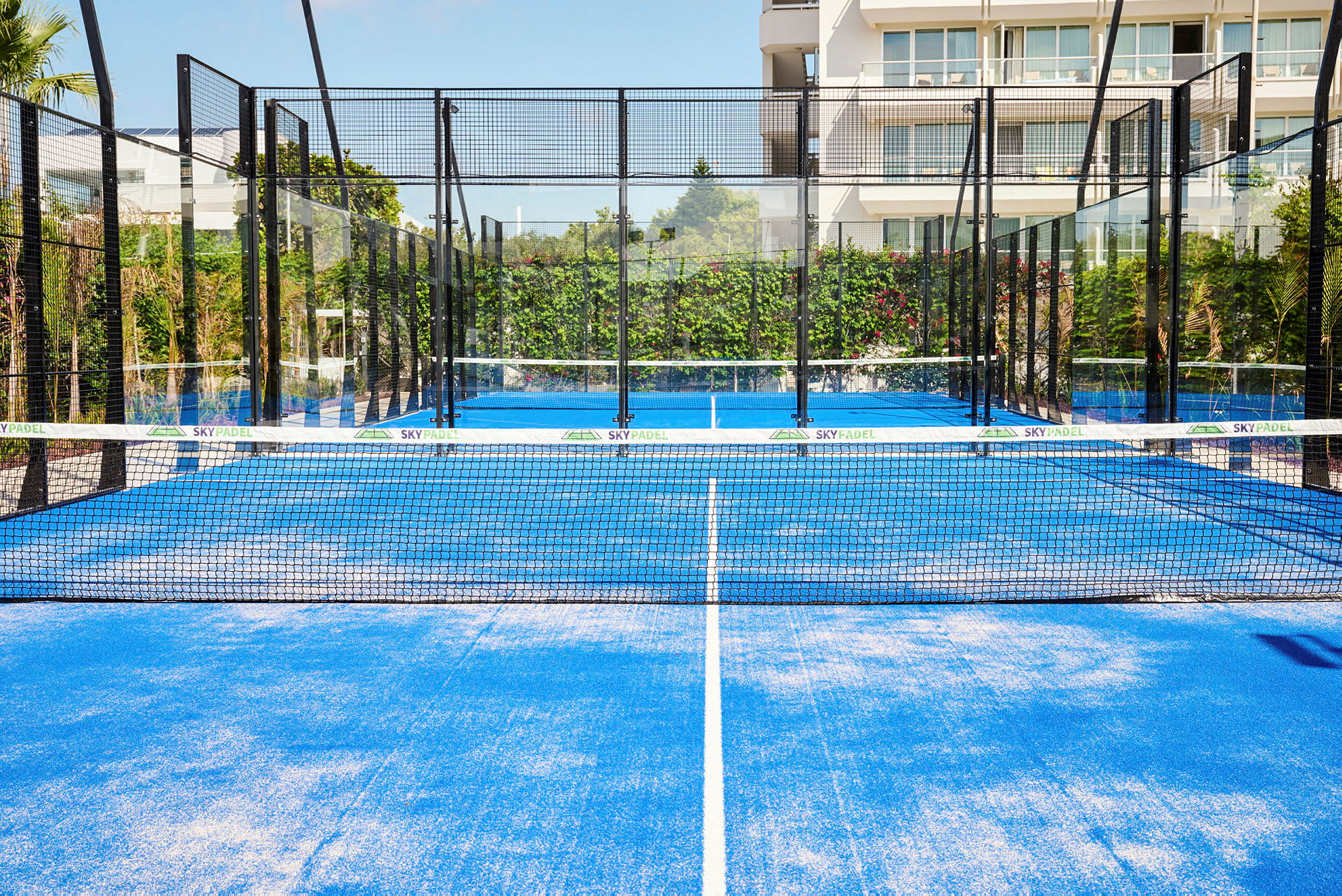 En blå padelbane med nett og omkringliggende glassvegger, plassert utendørs med vegetasjon og en bygning i bakgrunnen. (AI-generert tekst)