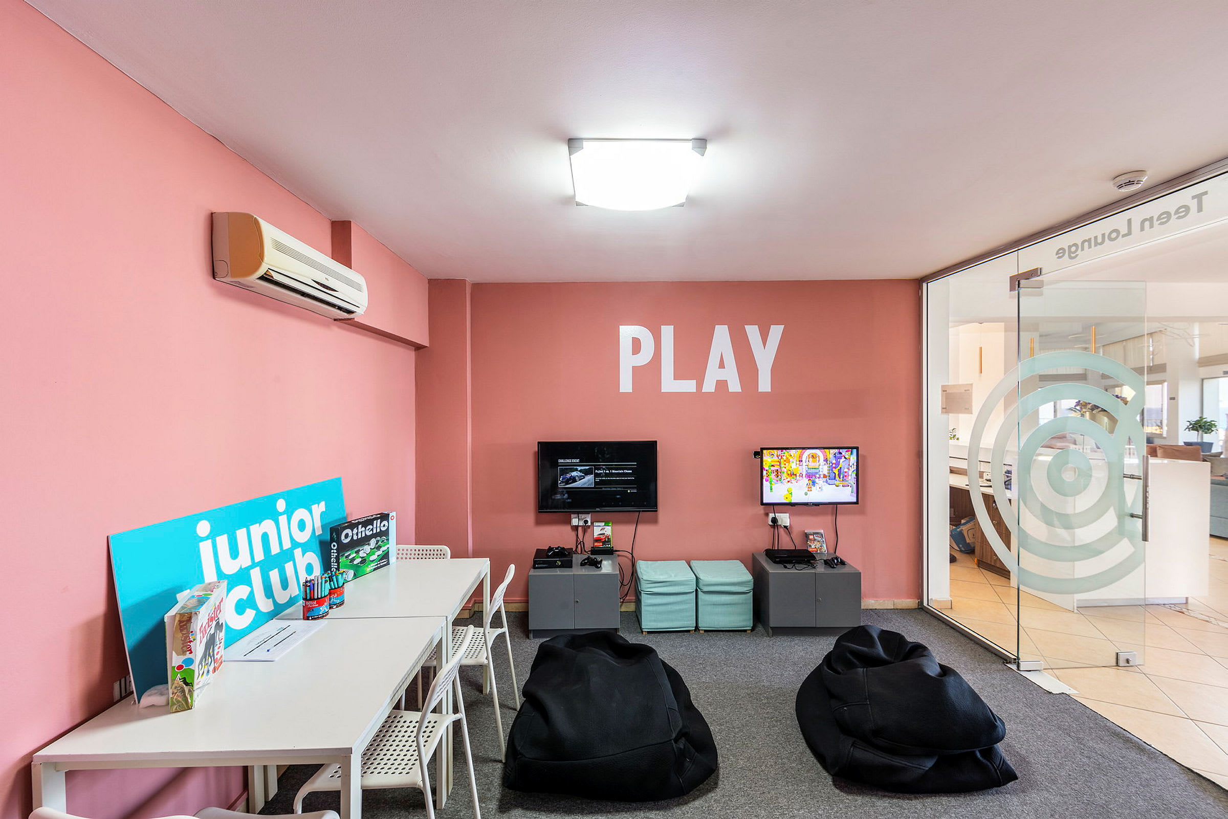 Et lekerom med rosa vegger, bord, stoler og spill samt sittesekker, og teksten 'PLAY' på veggen. (AI-generert tekst)