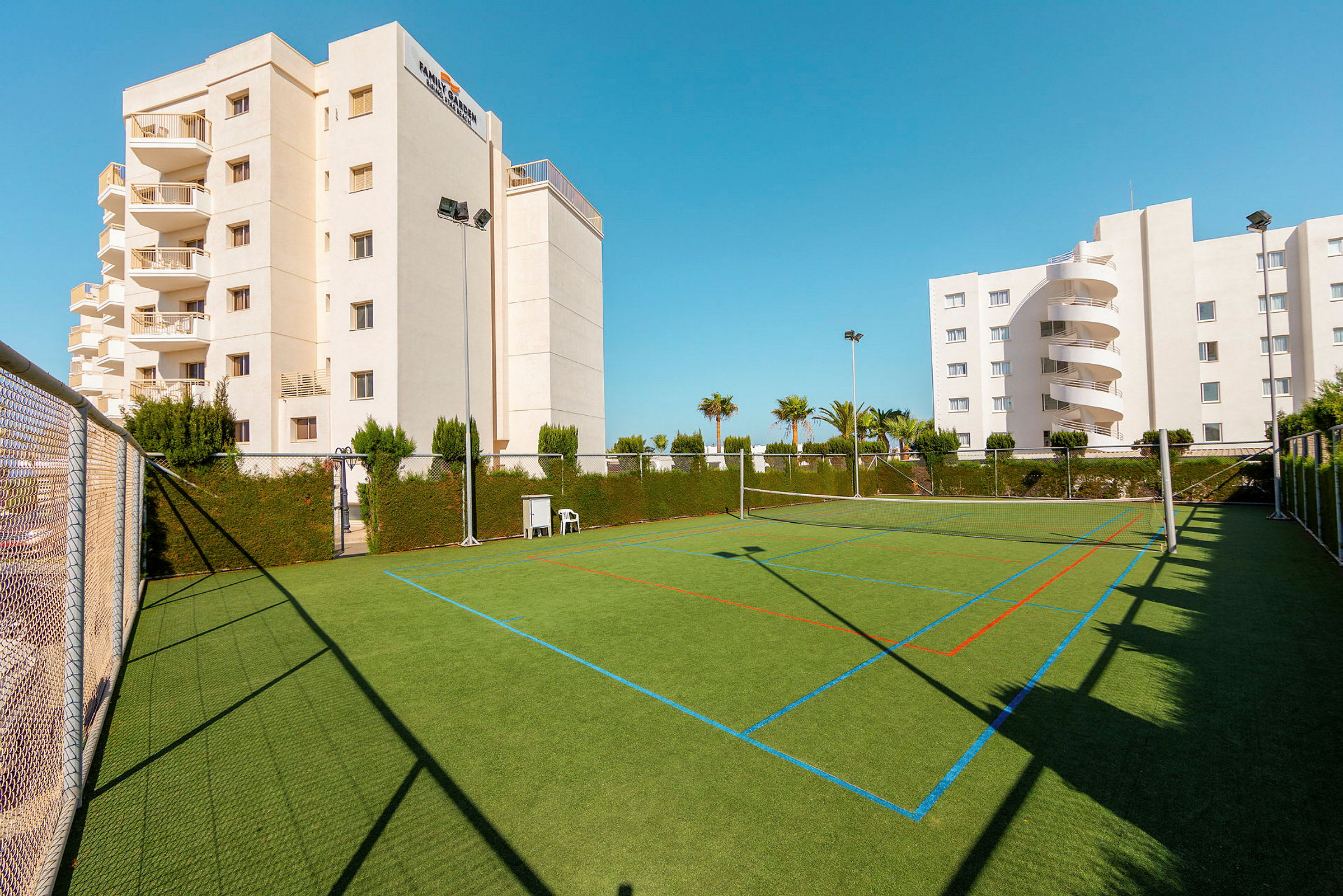 Fotballbane med grønt kunstgress, omgitt av gjerder og vegetasjon, ved siden av hotellbygninger. (AI-generert tekst)