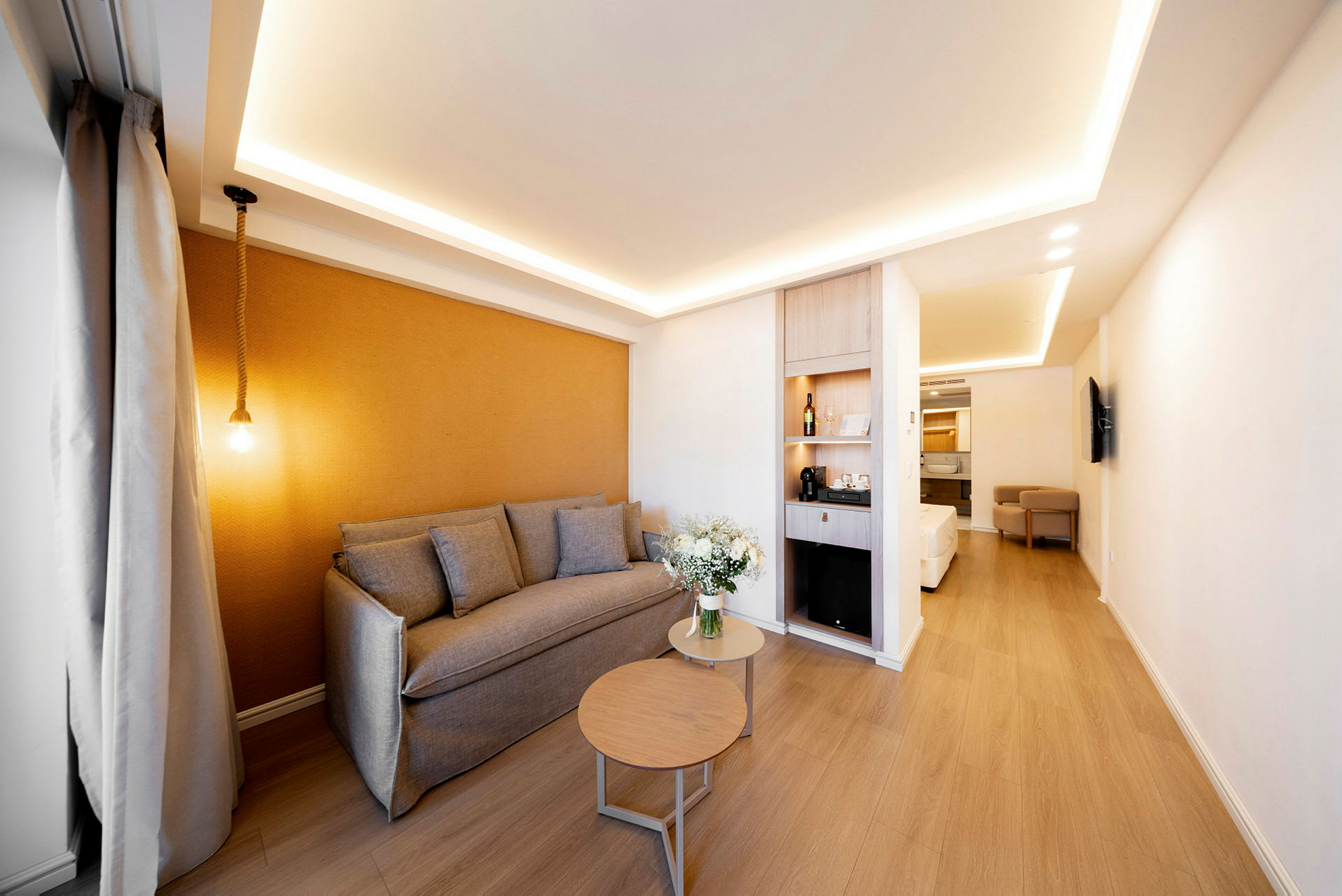 Et moderne hotellrom med lyse tredetaljer, en grå sofa og et rundt bord. En blomsterbukett står på bordet, og bakgrunnen har en brun aksentvegg. En minibar og en elegant seng ses lenger inn i rommet. (AI-generert tekst)