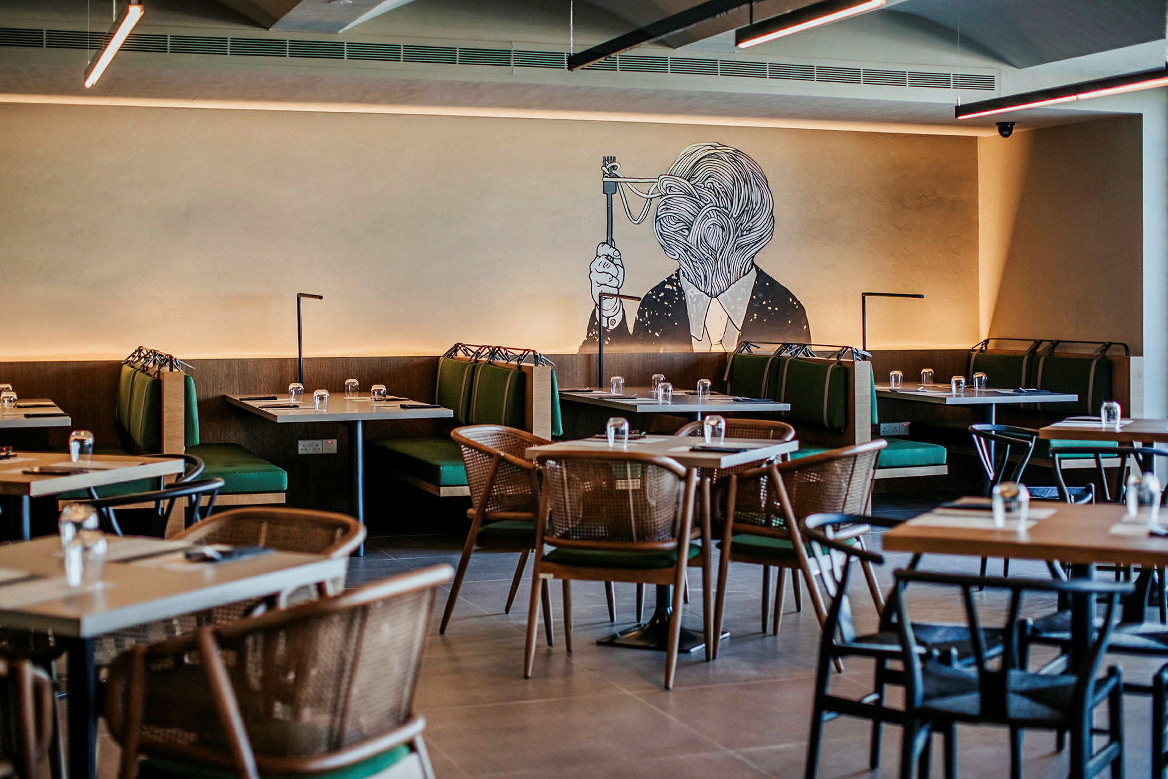 En restaurant med bord og grønne sofaer samt et veggmaleri av et kunstverk. (AI-generert tekst)