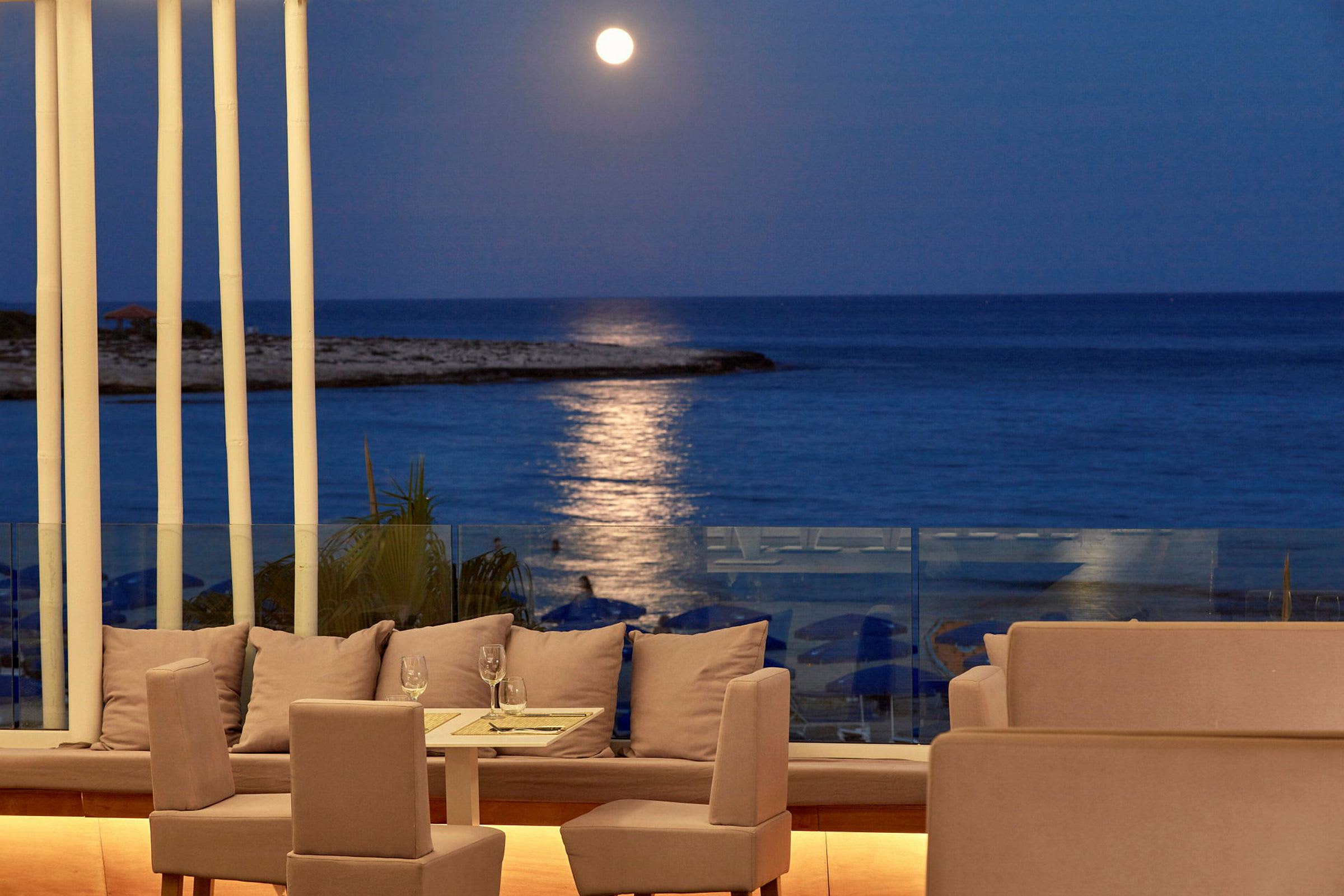 En terrasse med sitteplasser og et bord, med utsikt over havet under en klar månebelyst kveld. (AI-generert tekst)