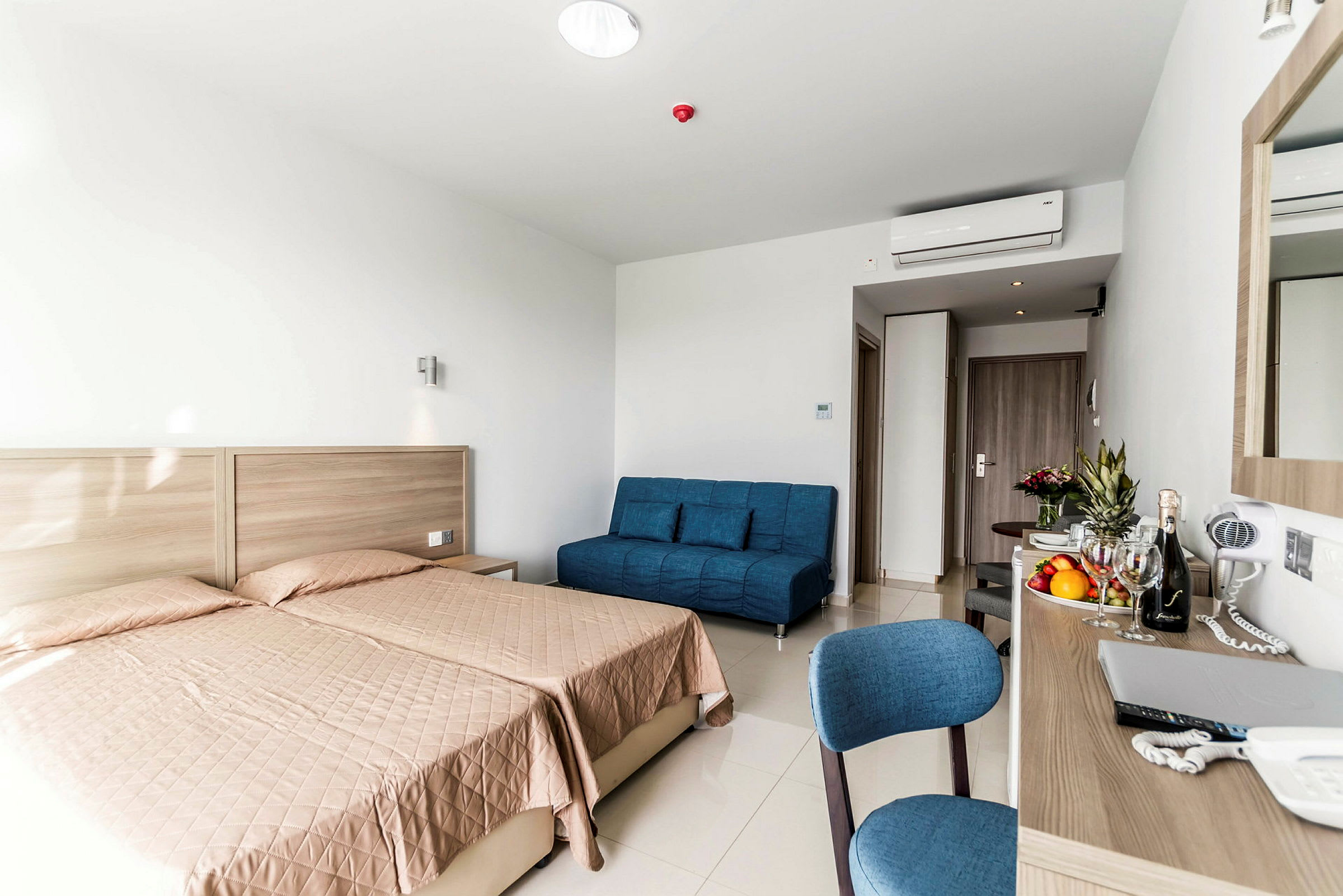 Et hotellrom med to enkeltsenger, en blå sofa og et skrivebord med diverse gjenstander. (AI-generert tekst)