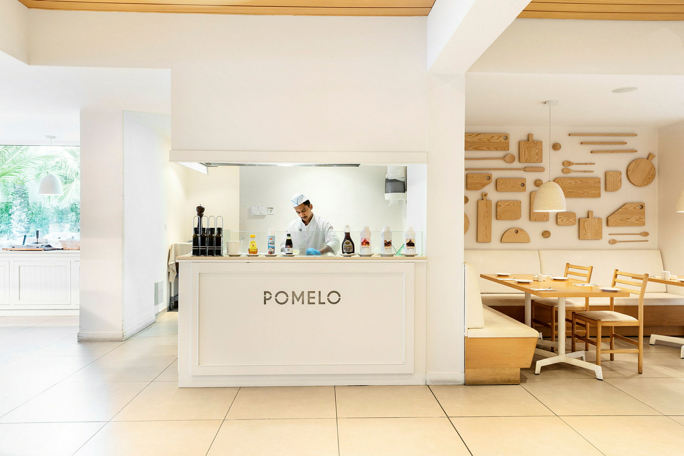 En restaurant med navnet 'Pomelo' og kjøkkenområde bak disken, møblert med lyse trebord og stoler samt dekorative trepaneler på veggen. (AI-generert tekst)