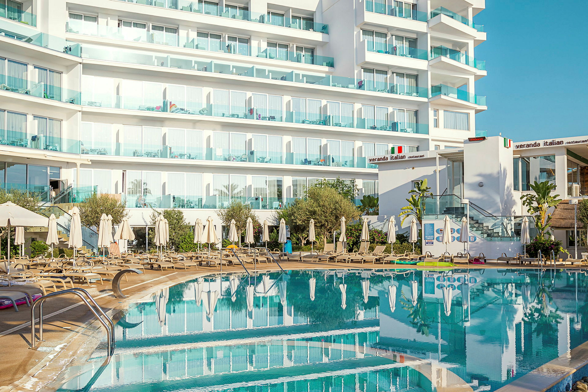Hotellbygning med balkonger bak et utendørsbasseng og solsenger. (AI-generert tekst)