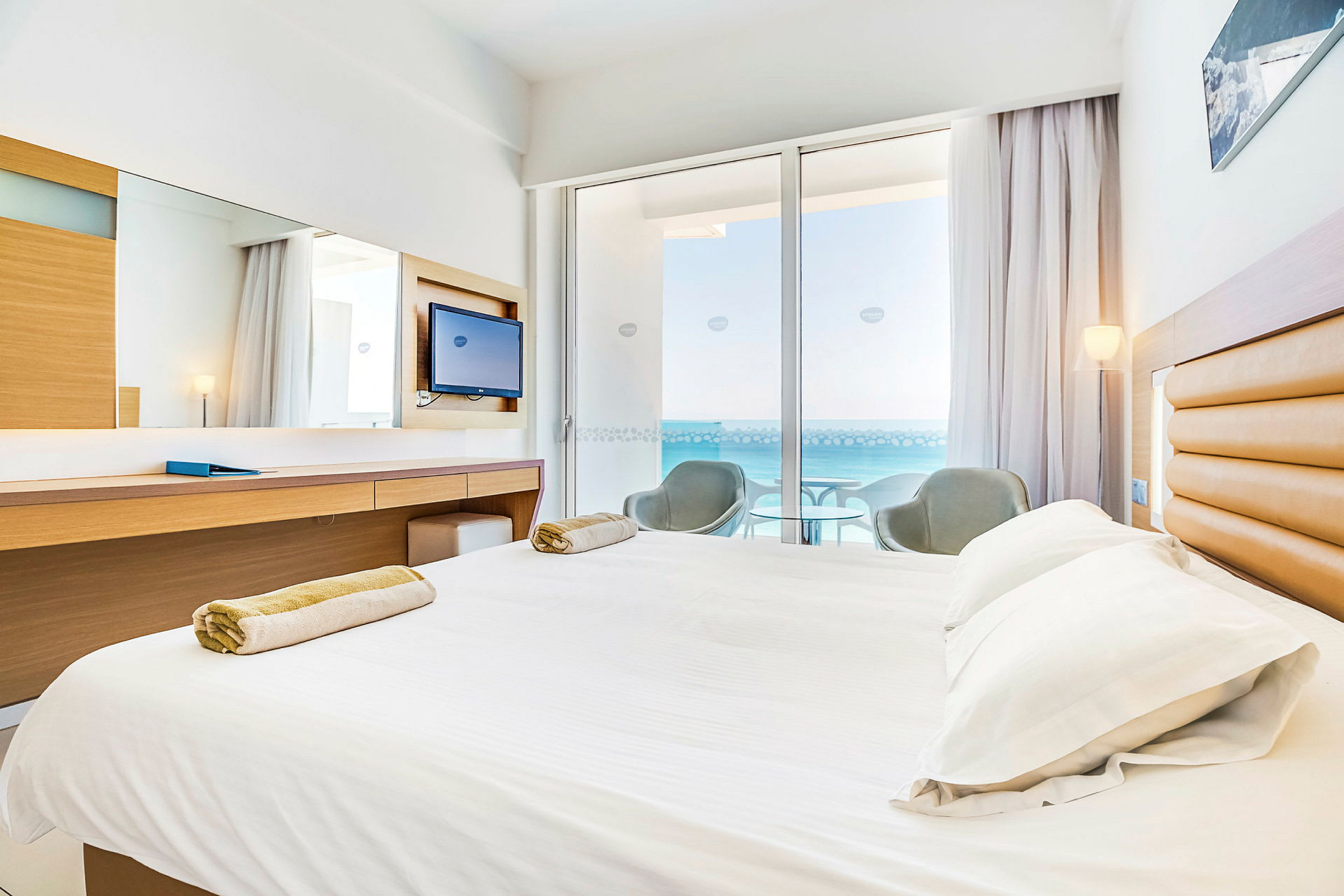 Hotellrom med seng, skrivebord og balkong med utsikt over havet. (AI-generert tekst)