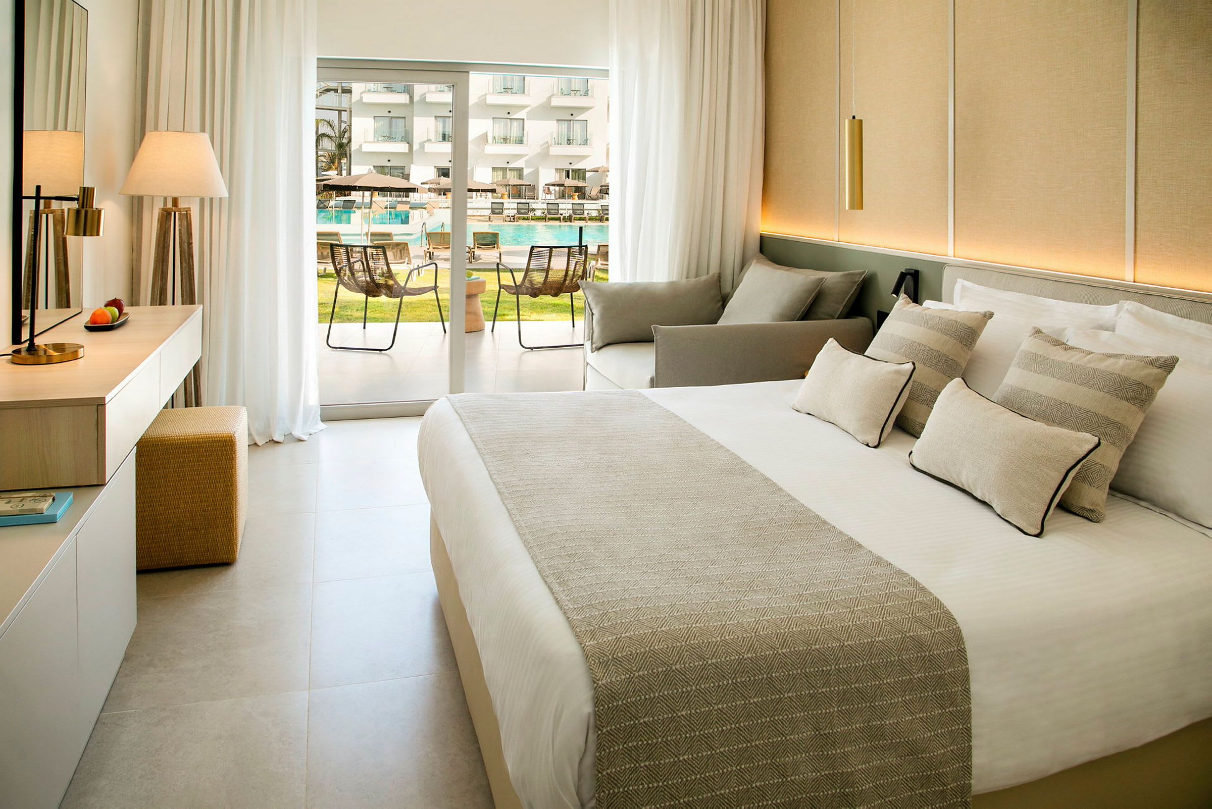 Moderne hotellrom med seng, sitteområde og balkongdør ut mot bassengområdet. (AI-generert tekst)