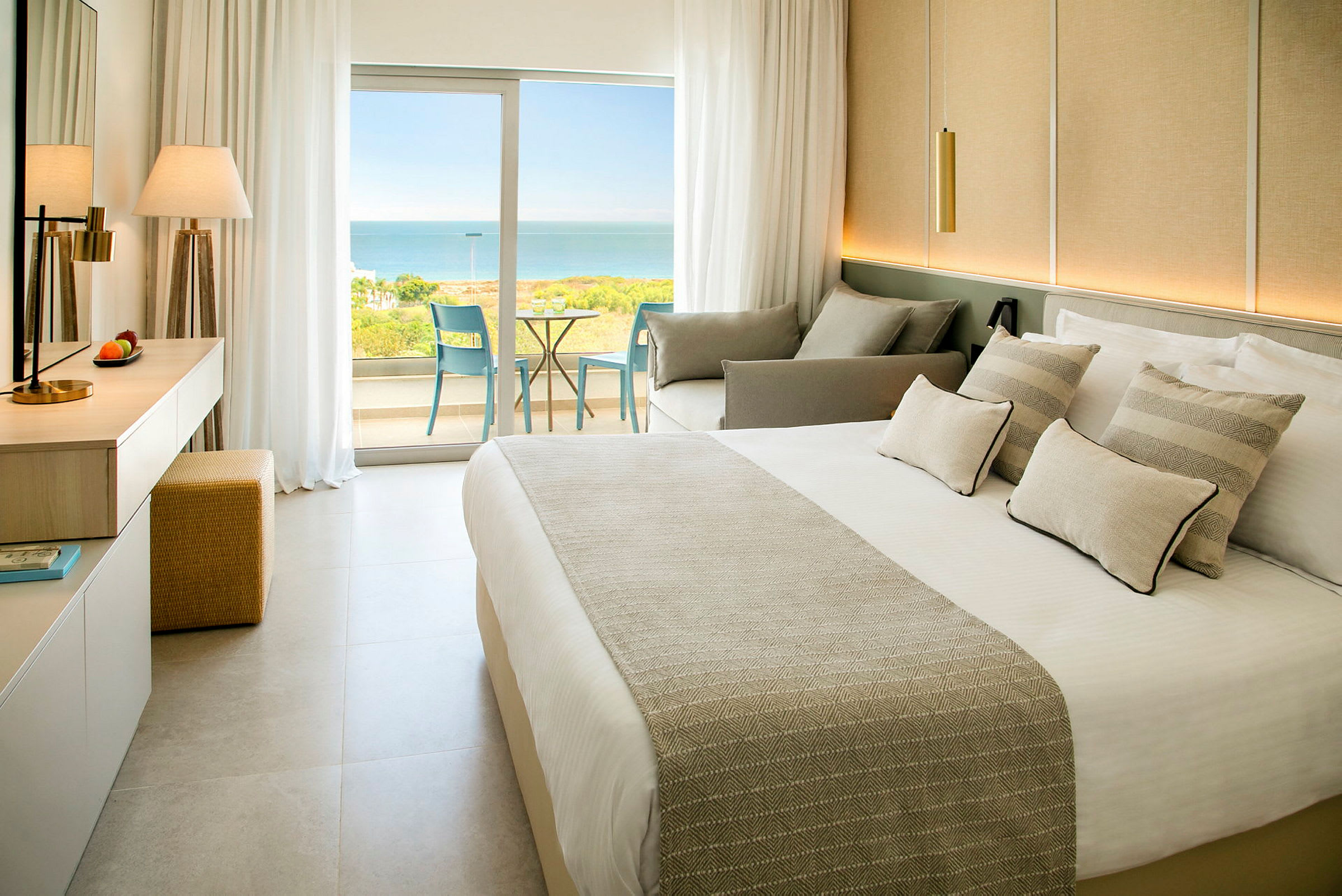 Et hotellrom med en seng, sofa og glassdører som leder ut til en balkong med utsikt over havet. (AI-generert tekst)