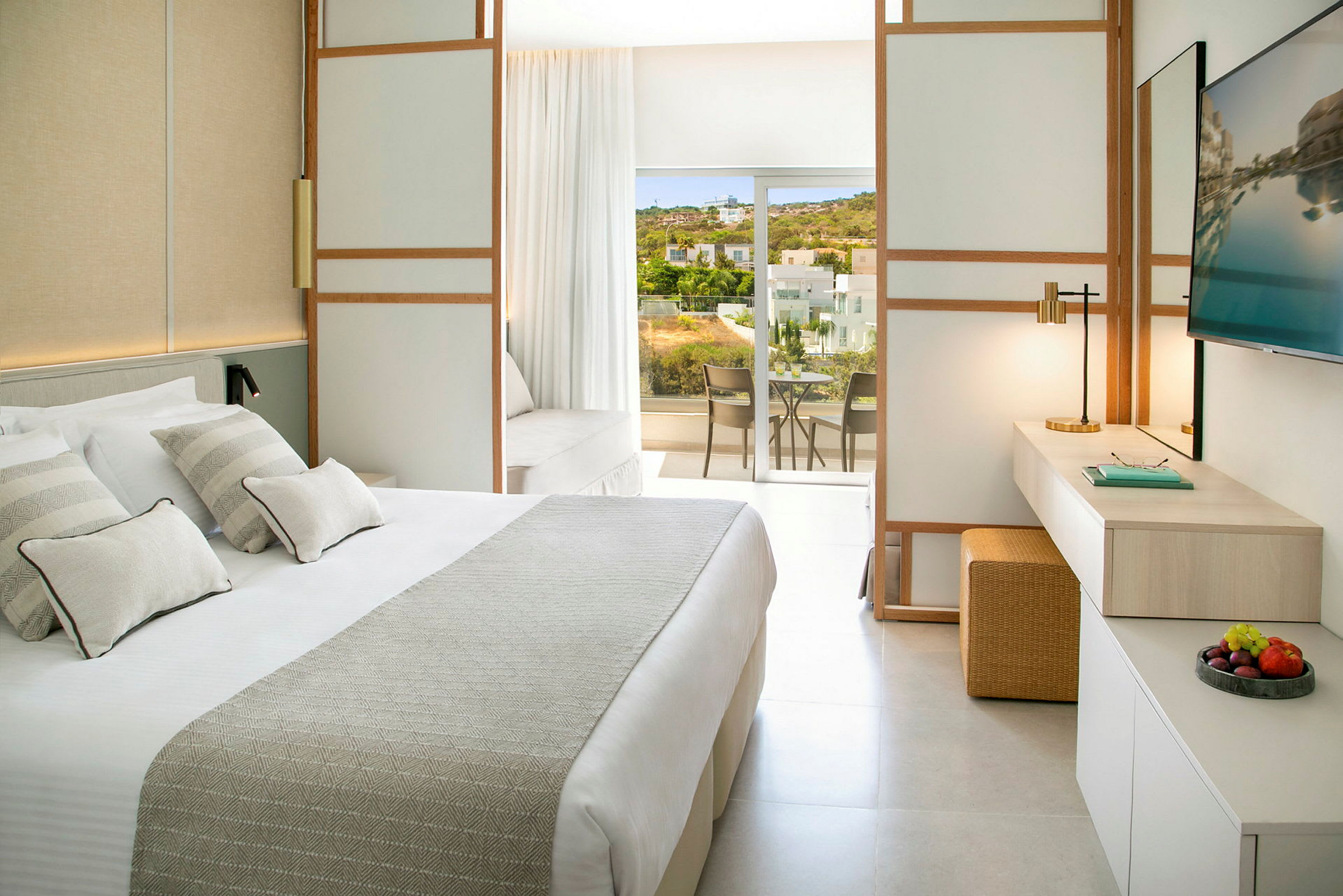 Et lyst hotellrom med en stor seng, en arbeidsplass og en balkong med utsikt over grøntarealer og bygninger. (AI-generert tekst)