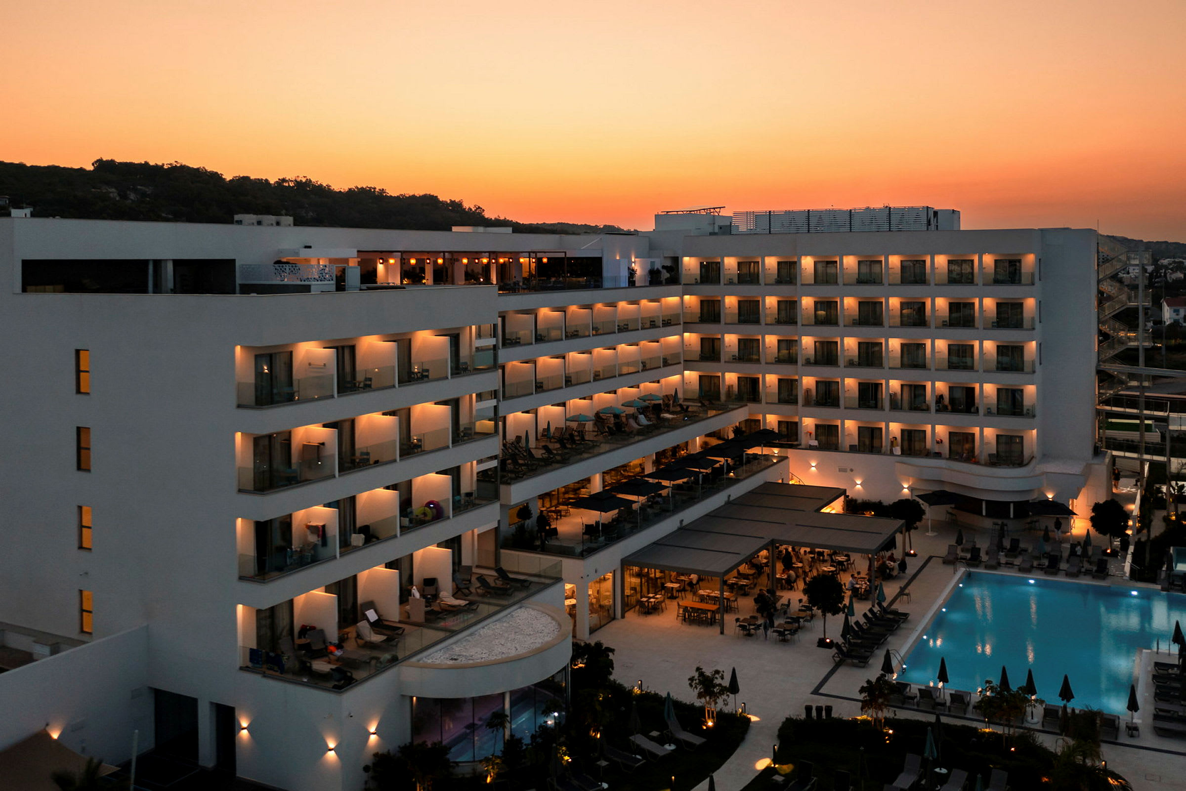 Moderne hotell med opplyste balkonger og et svømmebasseng ved solnedgang. (AI-generert tekst)