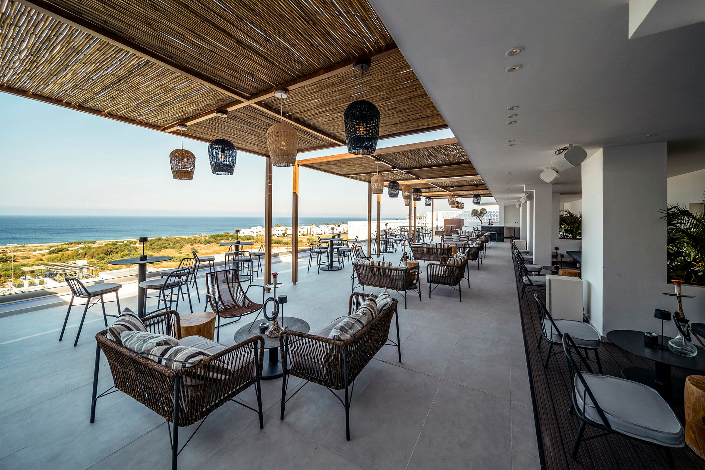 En terrasse med sitteplasser og tak av bambus, utsikt over havet og grønne områder. (AI-generert tekst)