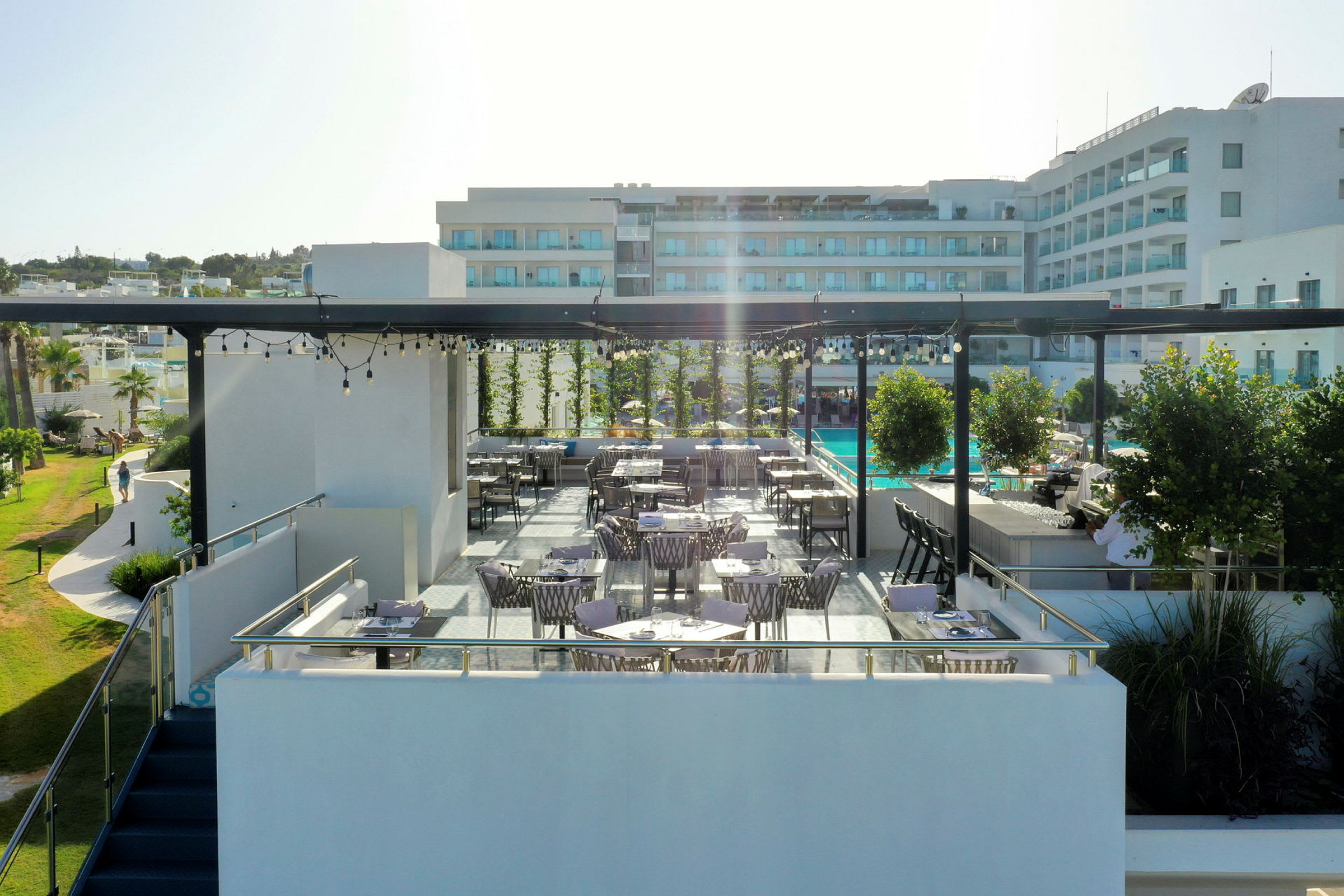 En utendørs terrasse med bord og stoler, omgitt av grønne planter og med en hotellbygning i bakgrunnen. (AI-generert tekst)