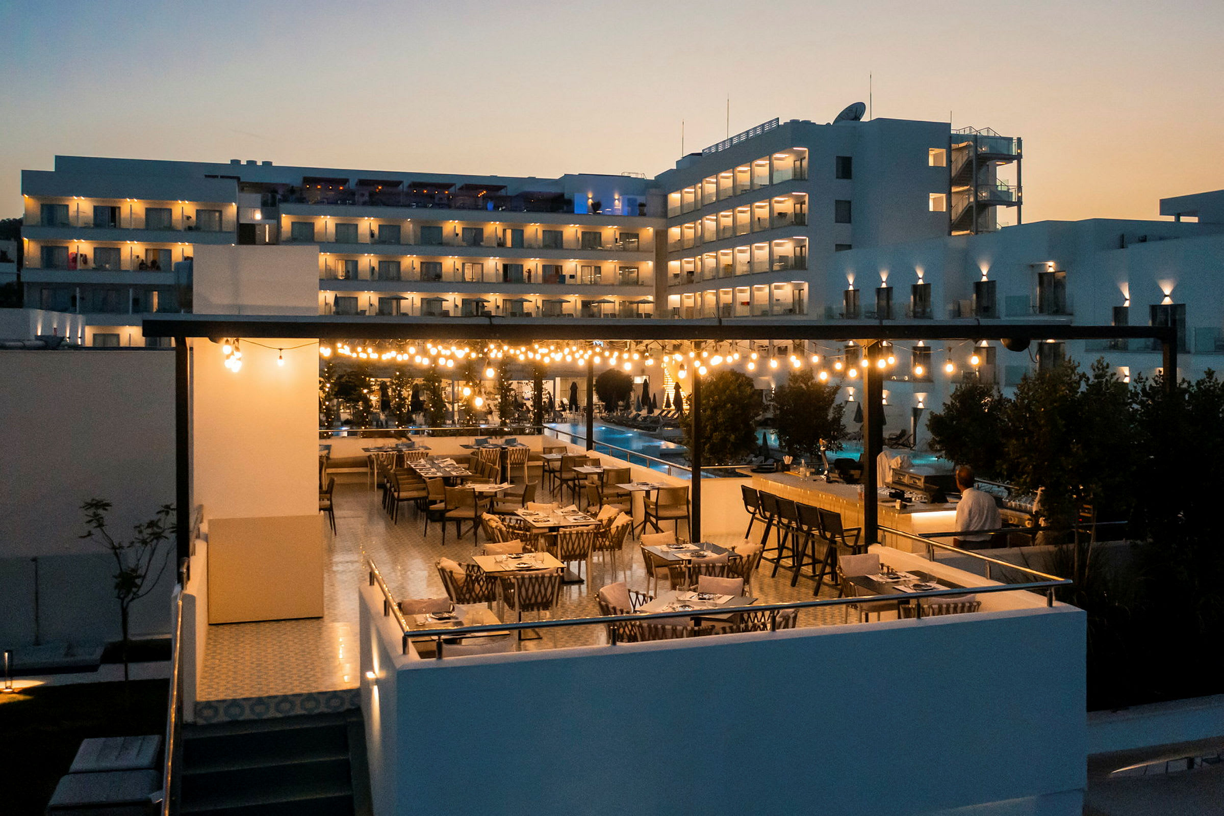 En restaurantterrasse med bord og stoler om kvelden, opplyst av lysslynger, med en moderne bygning i bakgrunnen. (AI-generert tekst)