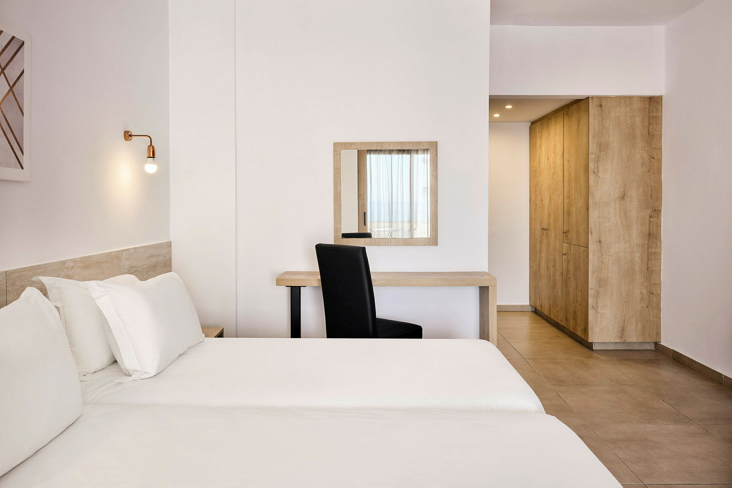Et lyst hotellrom med en seng, skrivebord, stol og garderobe. (AI-generert tekst)