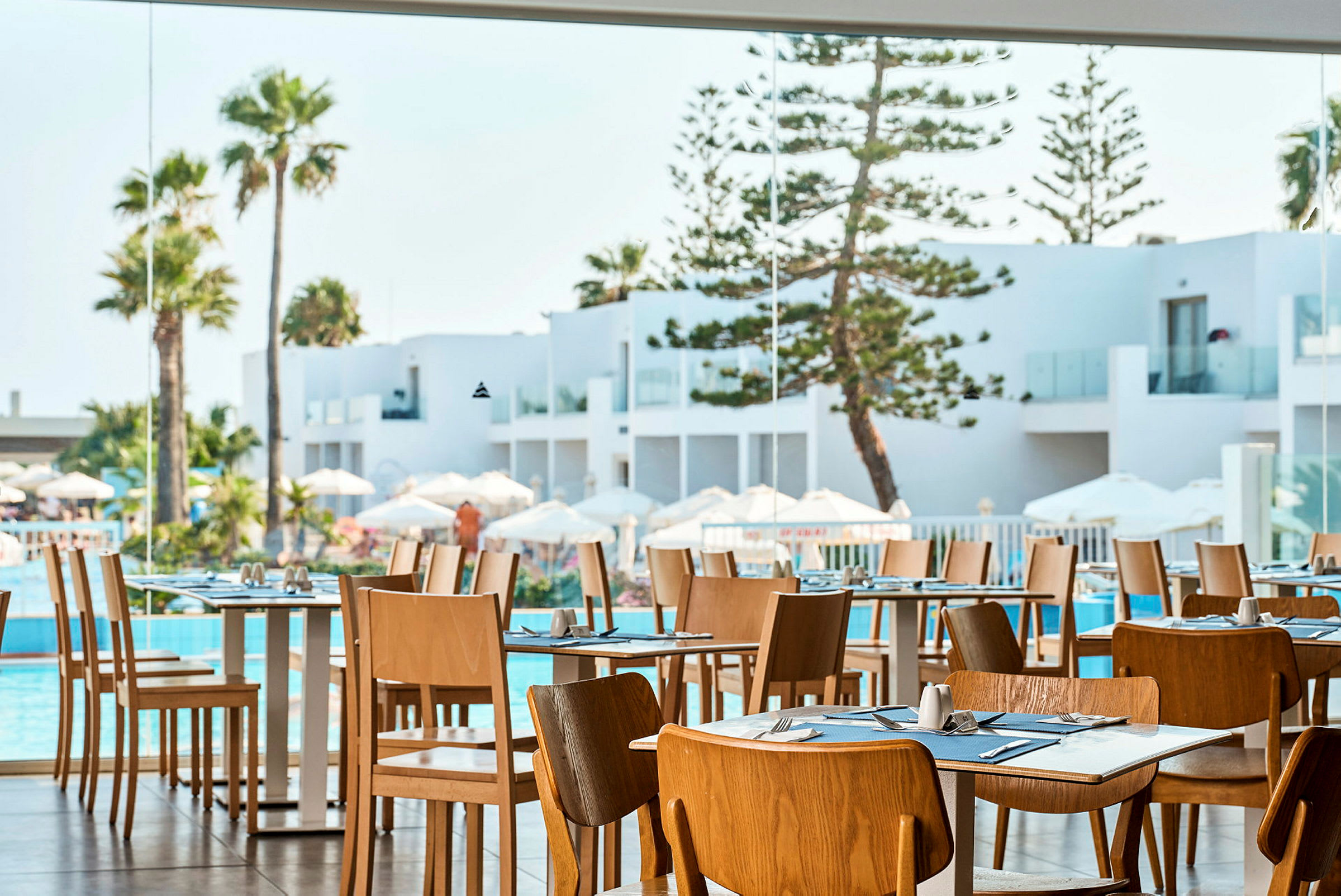 Restaurant med trestoler og dekte bord, utsikt mot hvit arkitektur, basseng og palmer. (AI-generert tekst)