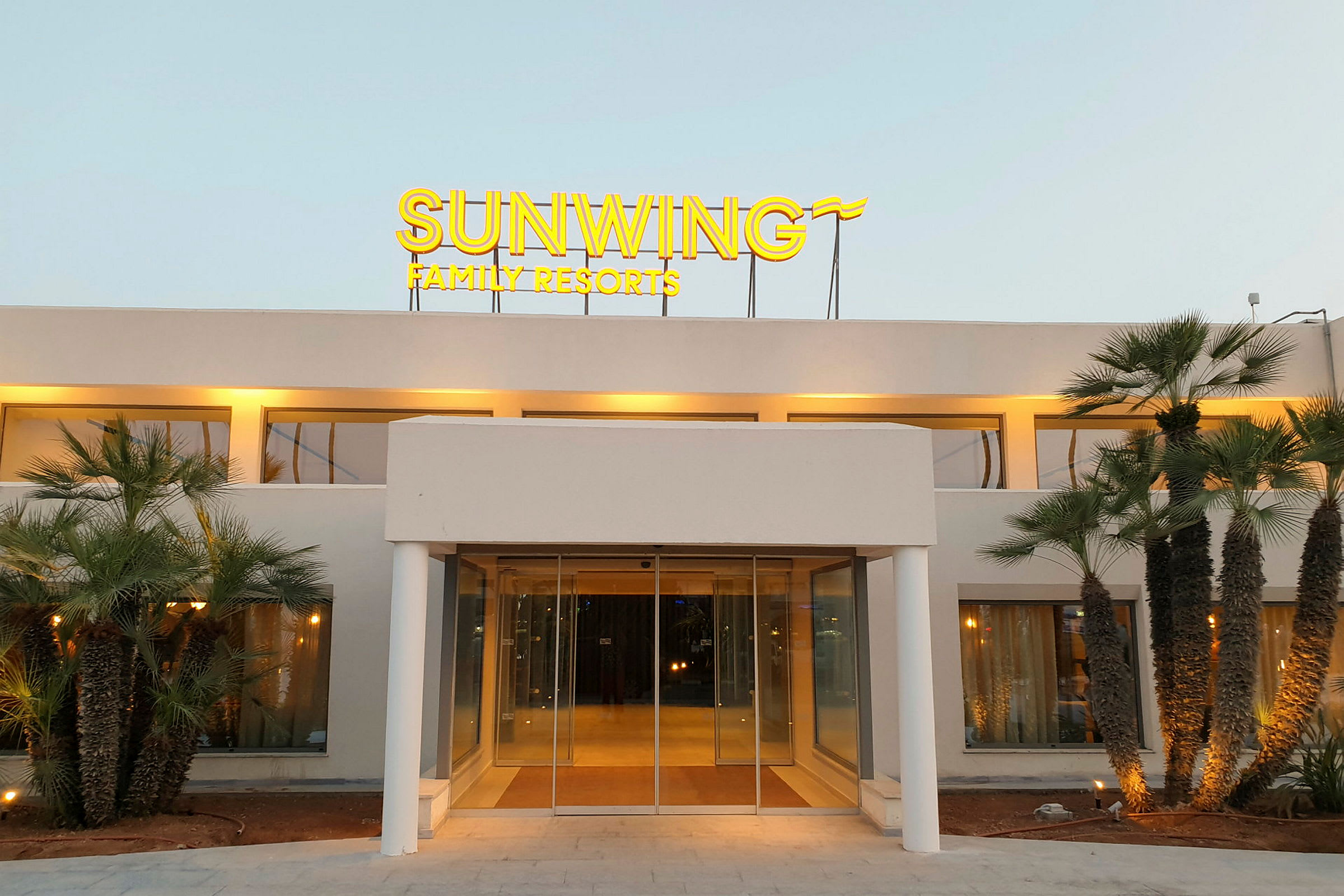 Forsiden av en bygning med et opplyst skilt som sier "Sunwing Family Resorts" og omgitt av palmer. (AI-generert tekst)