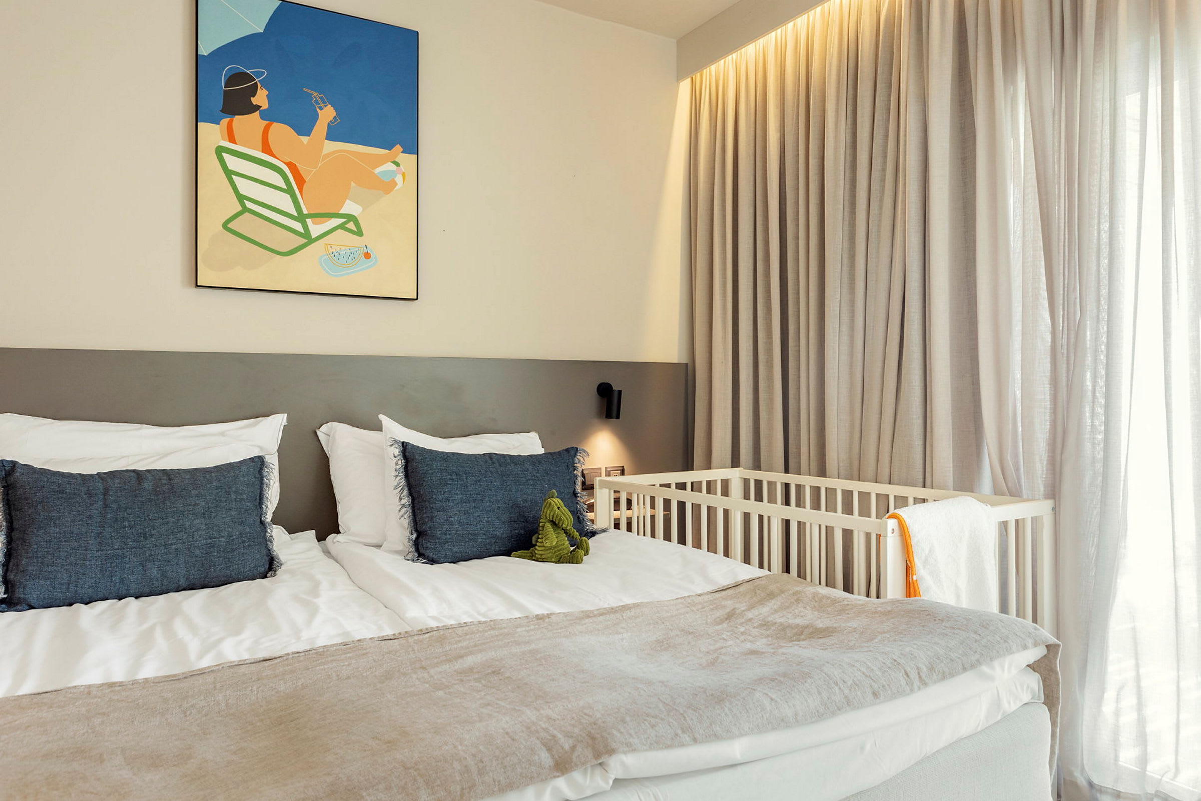 Et hotellrom med en dobbeltseng, pyntet med blå puter og en illustrasjon på veggen over. En babyseng er ved siden av vinduet med tynne gardiner. (AI-generert tekst)