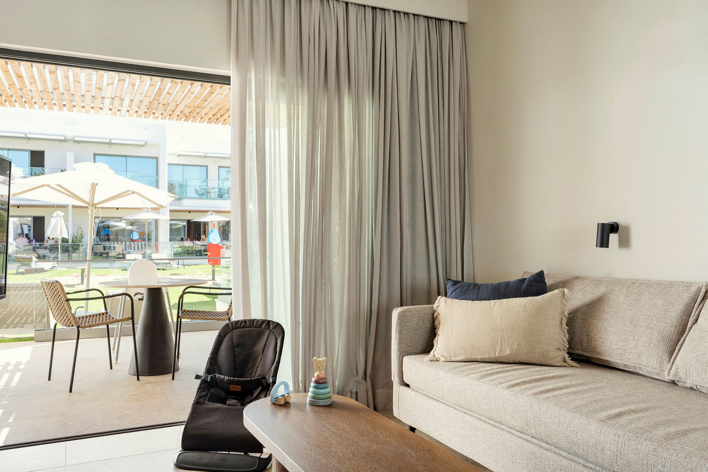Et moderne oppholdsrom med en sofa, et salongbord og en barnestol foran et stort vindu som vender ut mot en utendørsterrasse og hage. (AI-generert tekst)