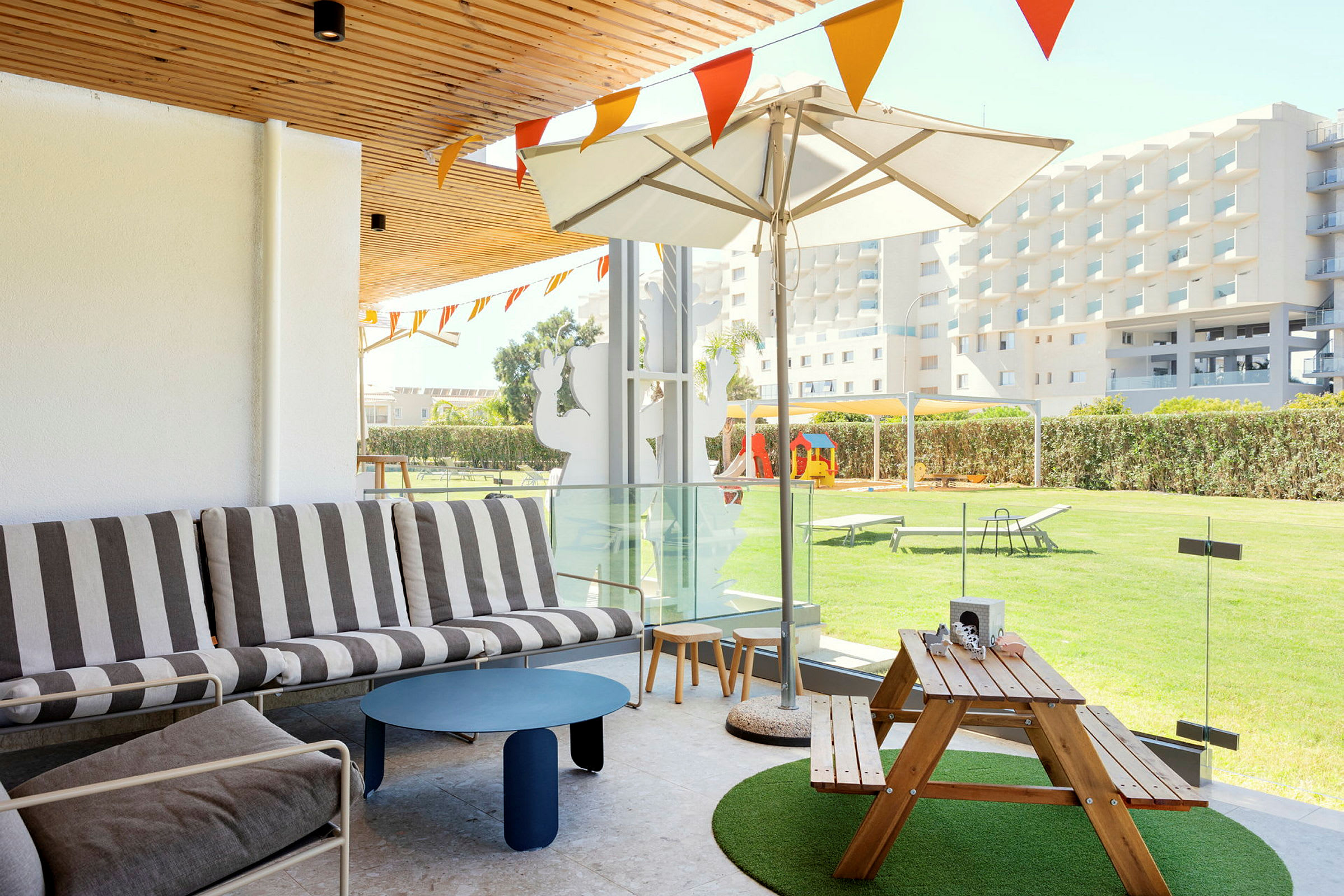 Utendørsmøbler med sitteområde og trebord på en terrasse, med utsikt over en plen og bygninger i bakgrunnen. (AI-generert tekst)