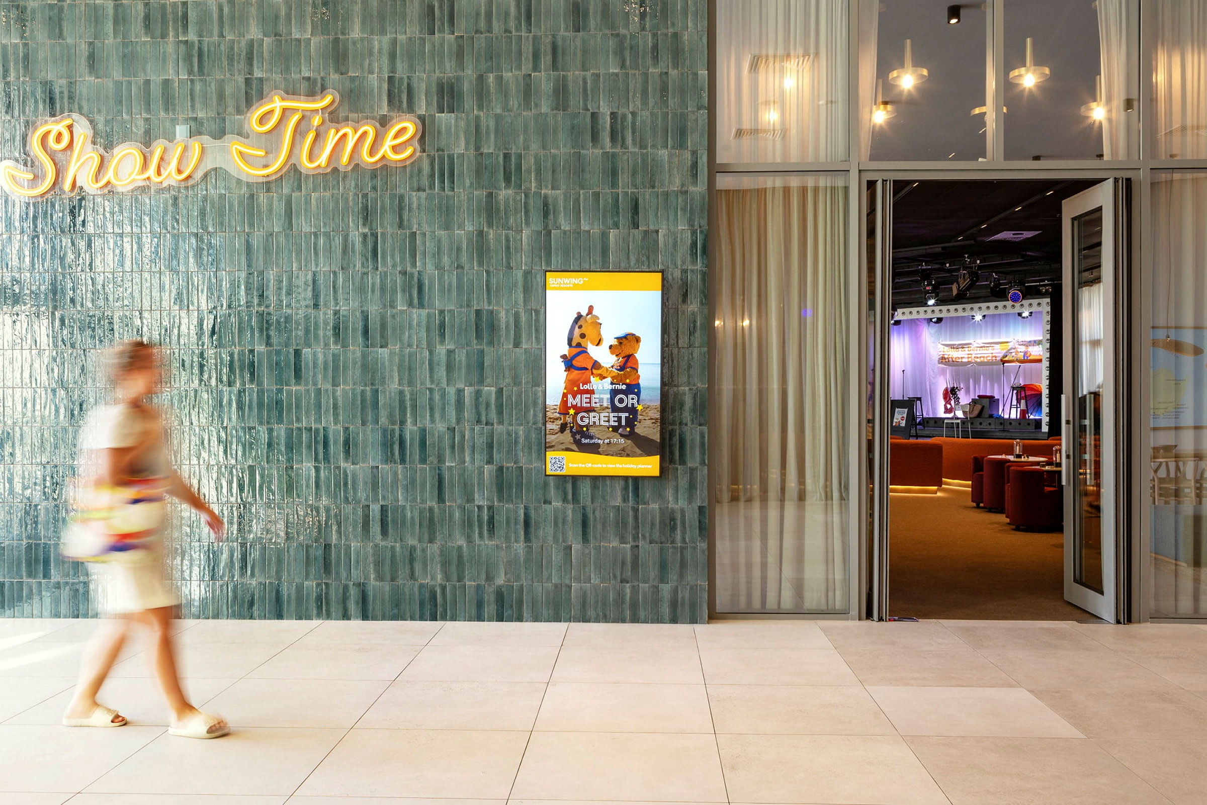 En person går forbi en vegg med teksten "Show Time" og en plakat for et barneprogram. I bakgrunnen vises en scene med publikumsstoler. (AI-generert tekst)