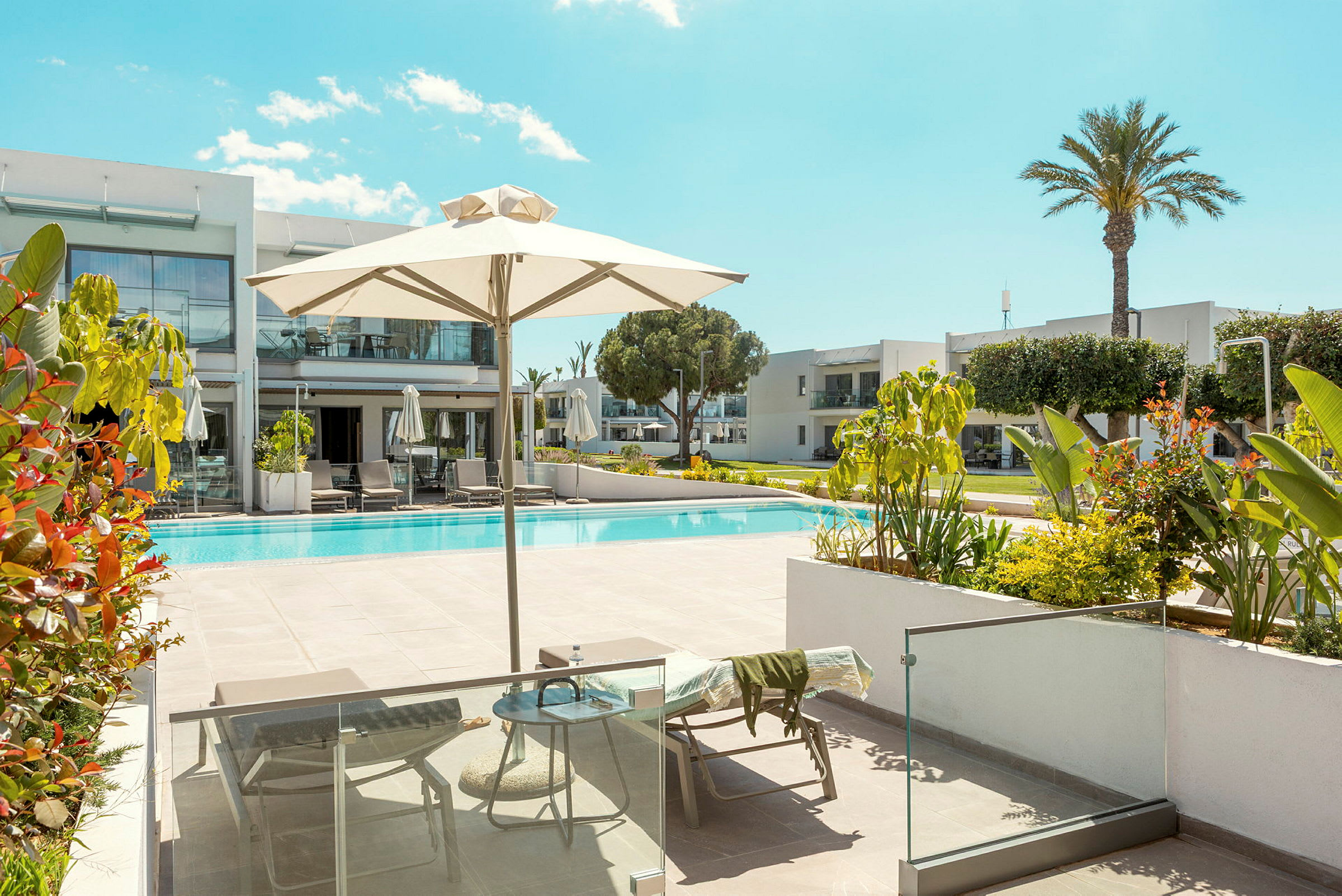 Utendørsmiljø med et basseng, solsenger og parasoller omgitt av moderne bygninger og grøntområder. (AI-generert tekst)