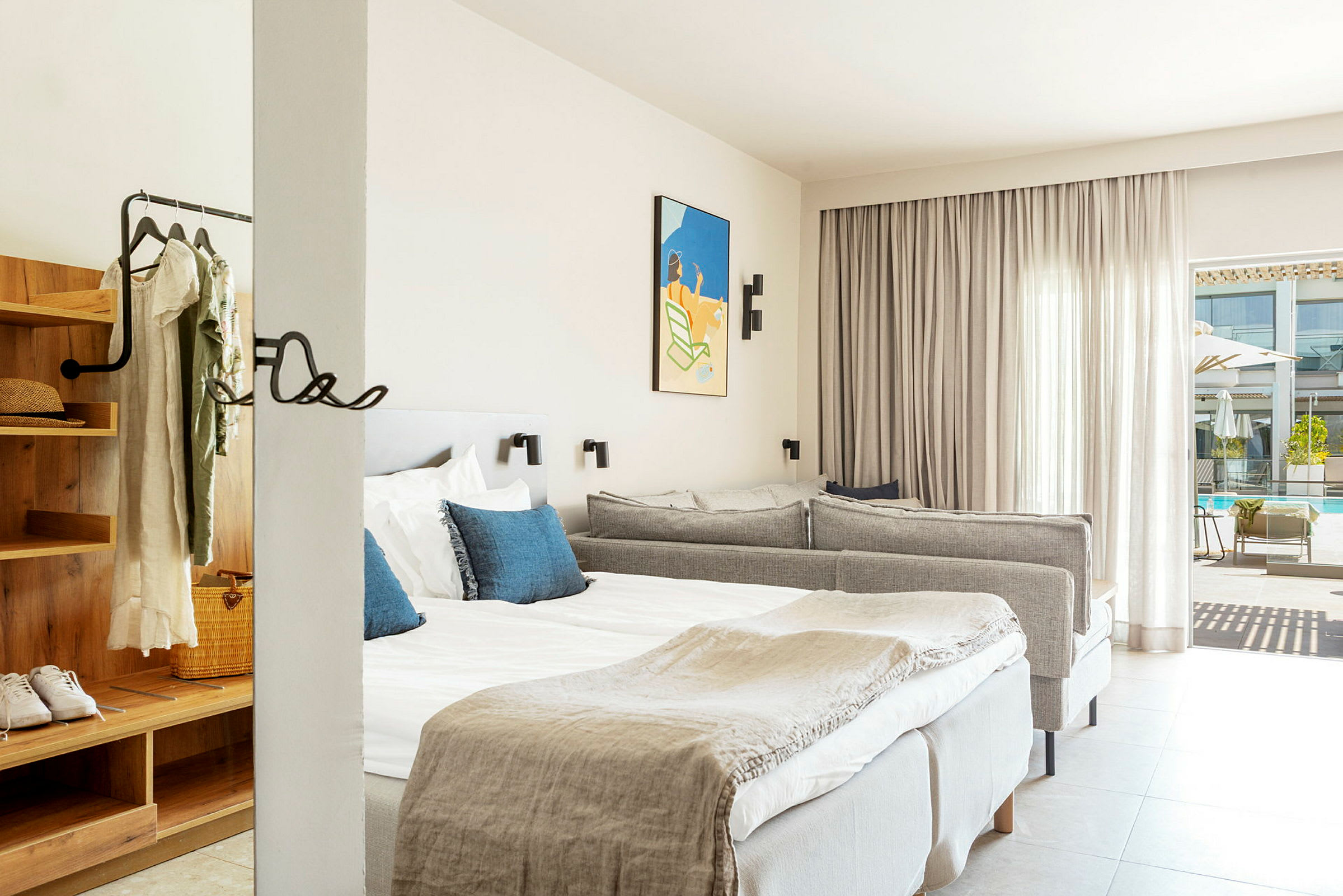 Et hotellrom med en dobbeltseng med hvite laken og blå dekorative puter, en grå sofa samt et åpent garderobeområde med klær hengende på kleshengere. Et kunstverk henger på veggen og en glassdør fører ut til et bassengområde. (AI-generert tekst)