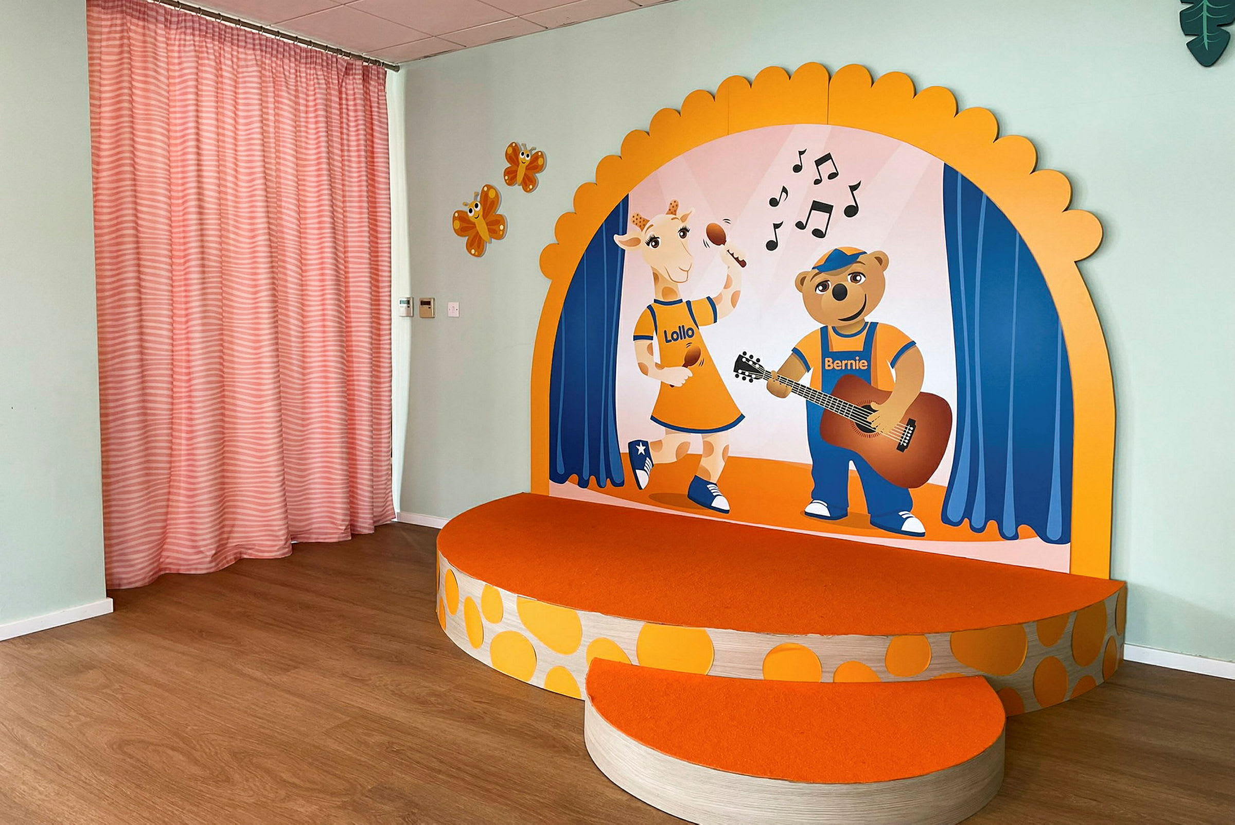 En liten opphøyet podium med oransje overflate og illustrasjoner av en giraff og en bjørn på en bakgrunn med musikkscene-tema. På sideveggen er oransje figurer og rosa gardiner. (AI-generert tekst)