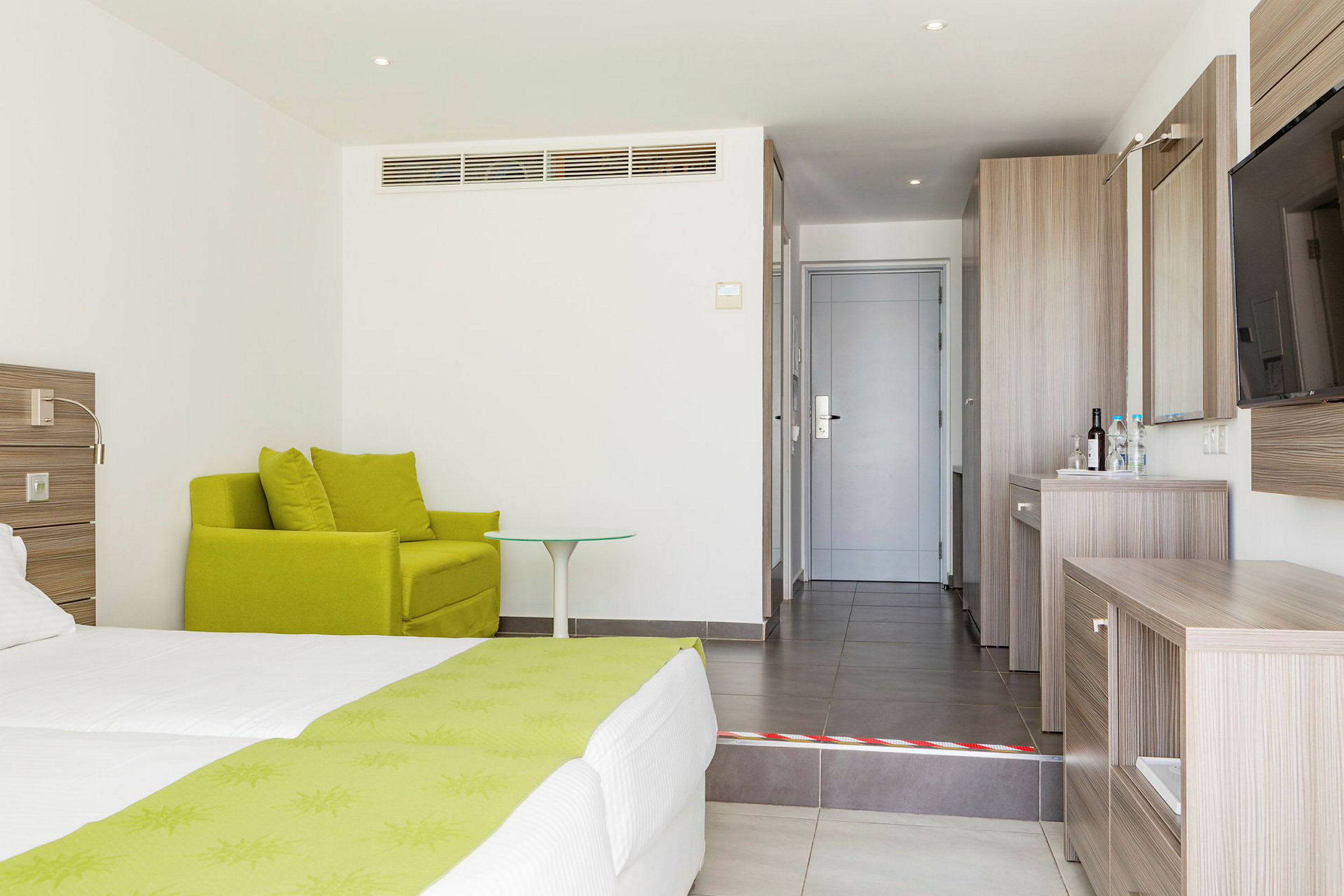 Hotellrom med grønn lenestol, seng med grønne detaljer og skrivebord i tre ved inngangen. (AI-generert tekst)
