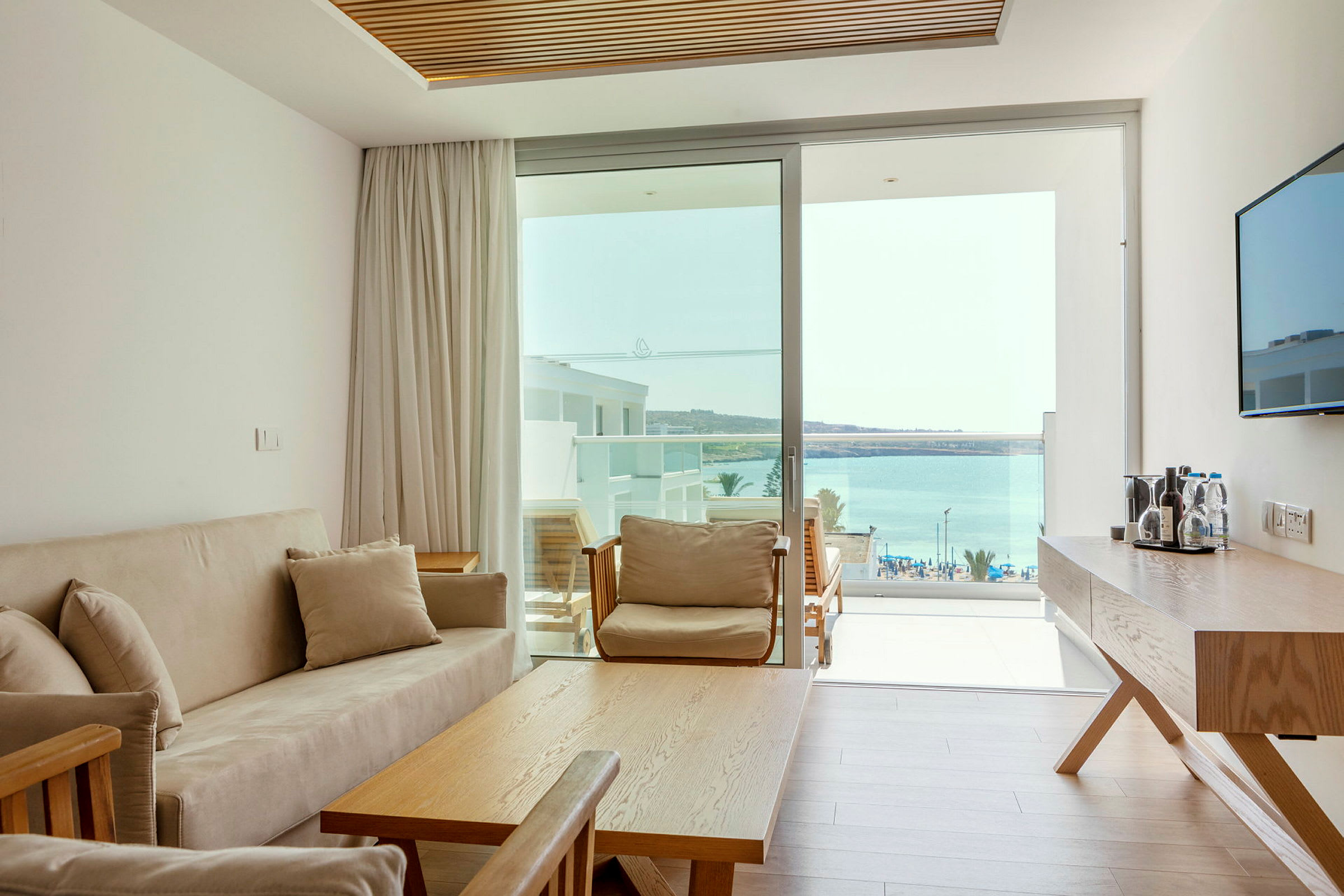 Et lyst hotellrom med en sofa, bord og utsikt over havet gjennom glassdører. (AI-generert tekst)