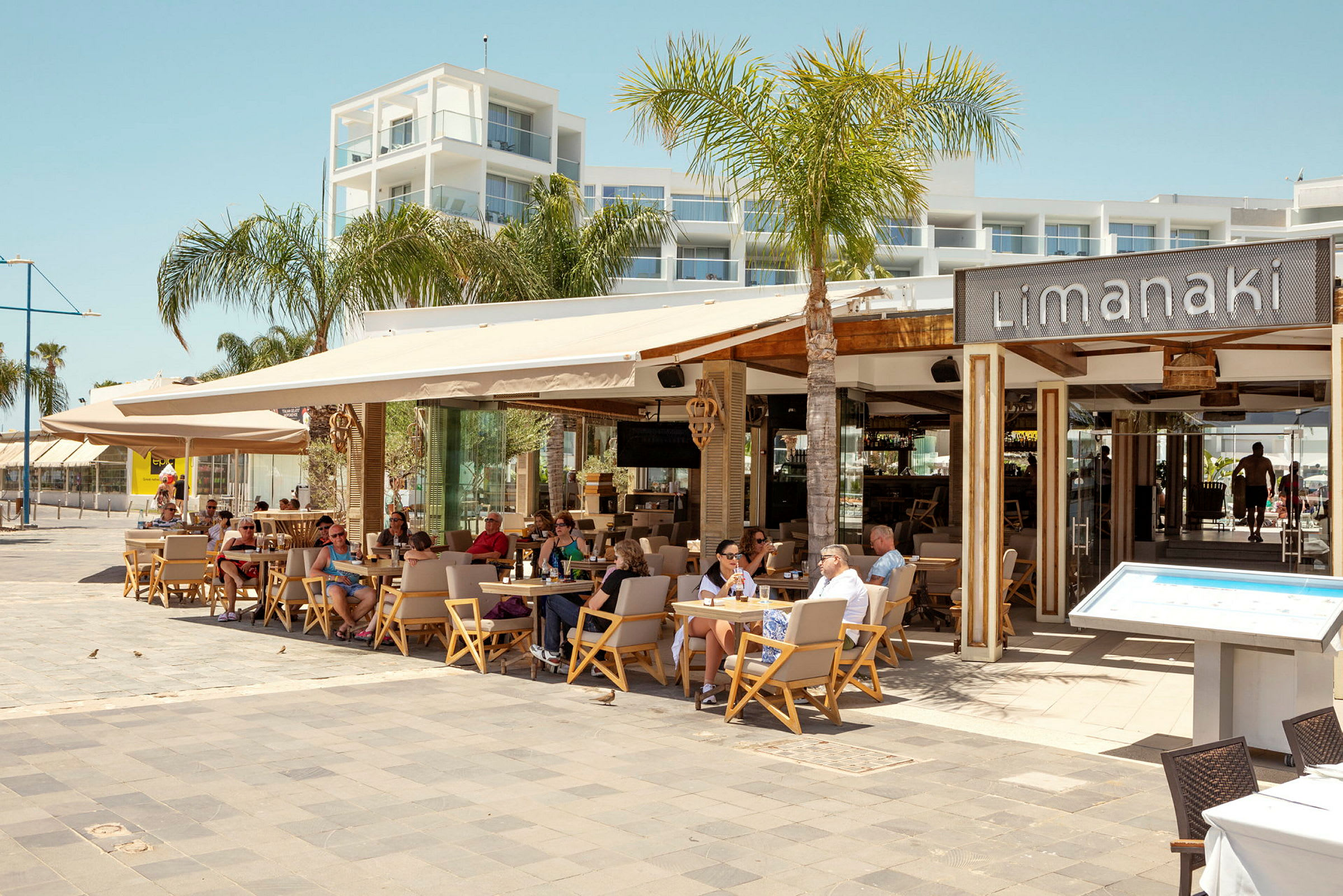 Restaurant Limanaki med sittende gjester, omgitt av palmer og moderne arkitektur. (AI-generert tekst)