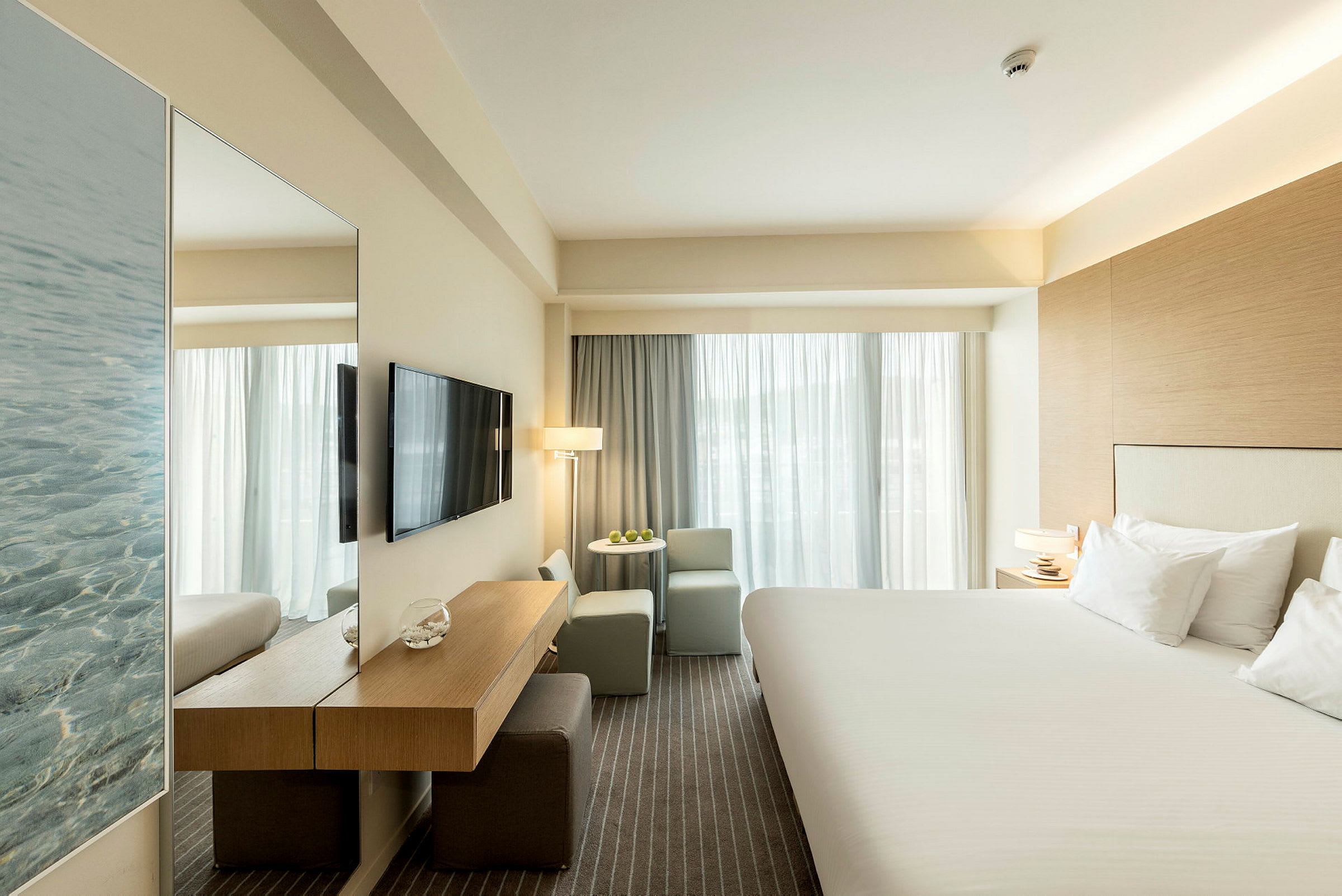 Hotellrom med en stor seng, TV på veggen og sittegruppe ved vinduet. (AI-generert tekst)