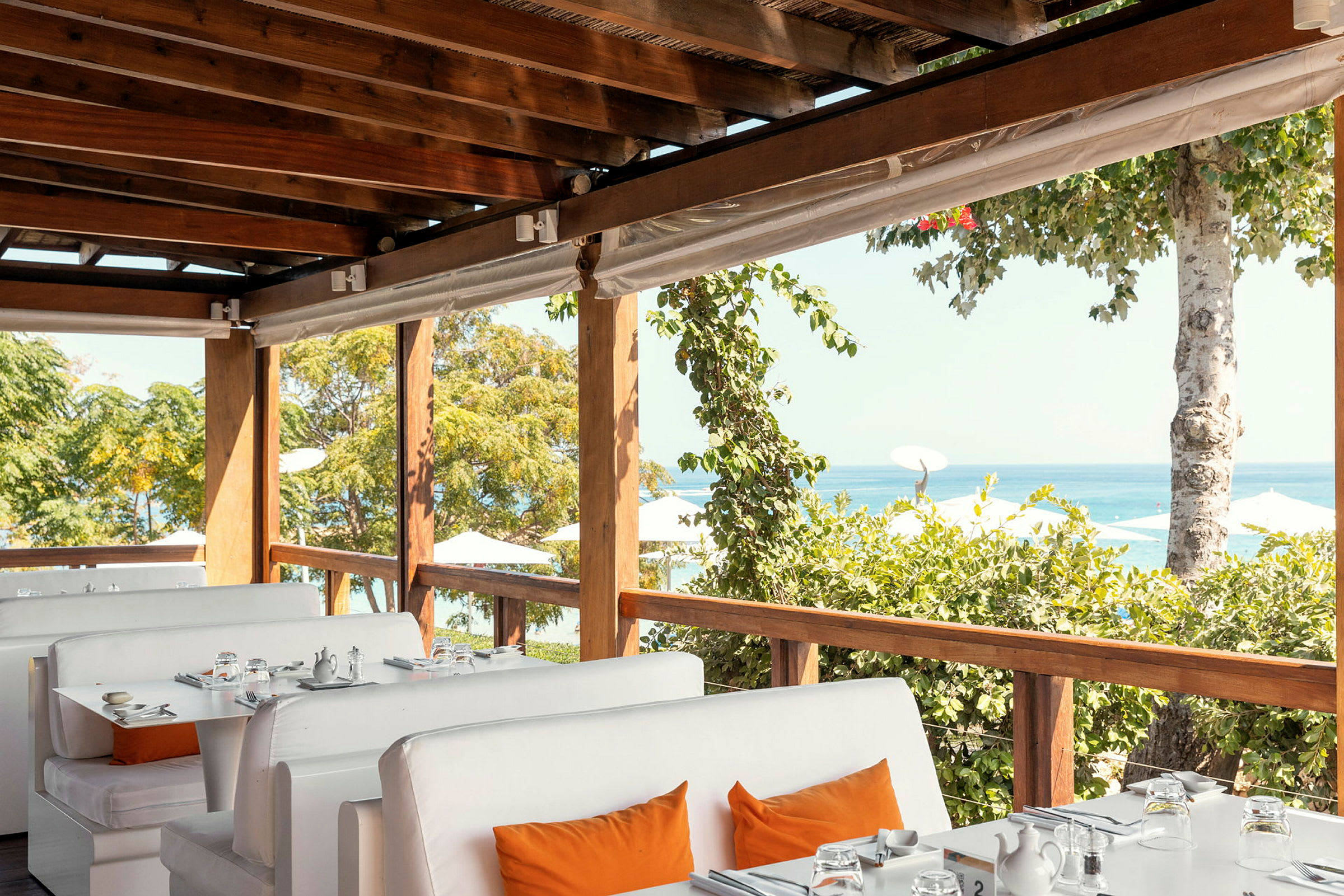 Bilde av en restaurant med hvite sofaer og bord med utsikt over havet og vegetasjonen. (AI-generert tekst)