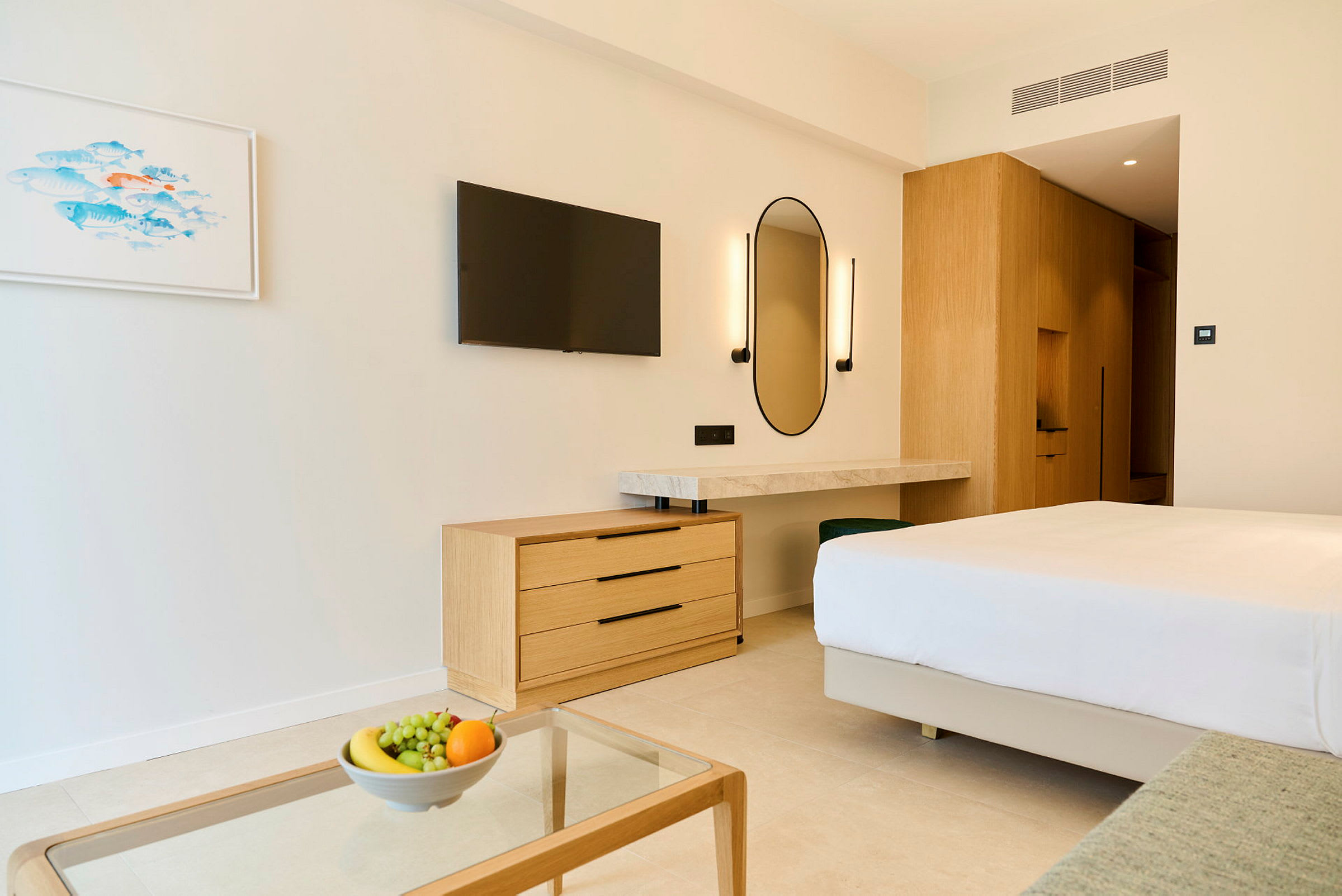 Et moderne hotellrom med en dobbeltseng, en kommode, en veggmontert TV og et skrivebord med speil. På et glassbord står en bolle med frukt. (AI-generert tekst)