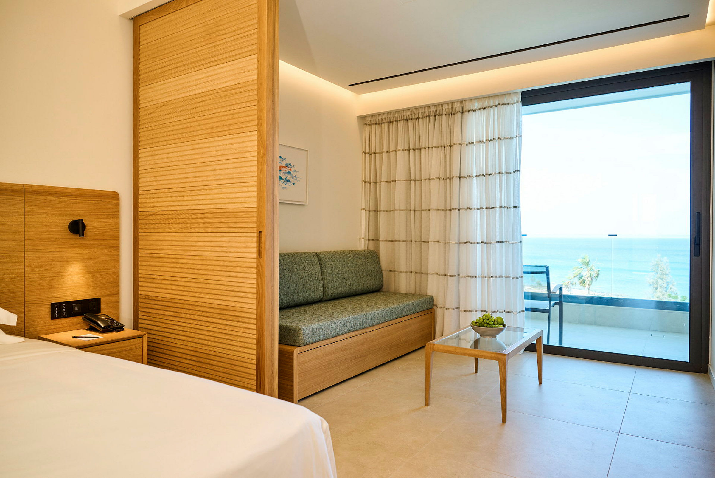 Et hotellrom med moderne innredning, en sofa, et lite bord og en balkong med havutsikt. (AI-generert tekst)