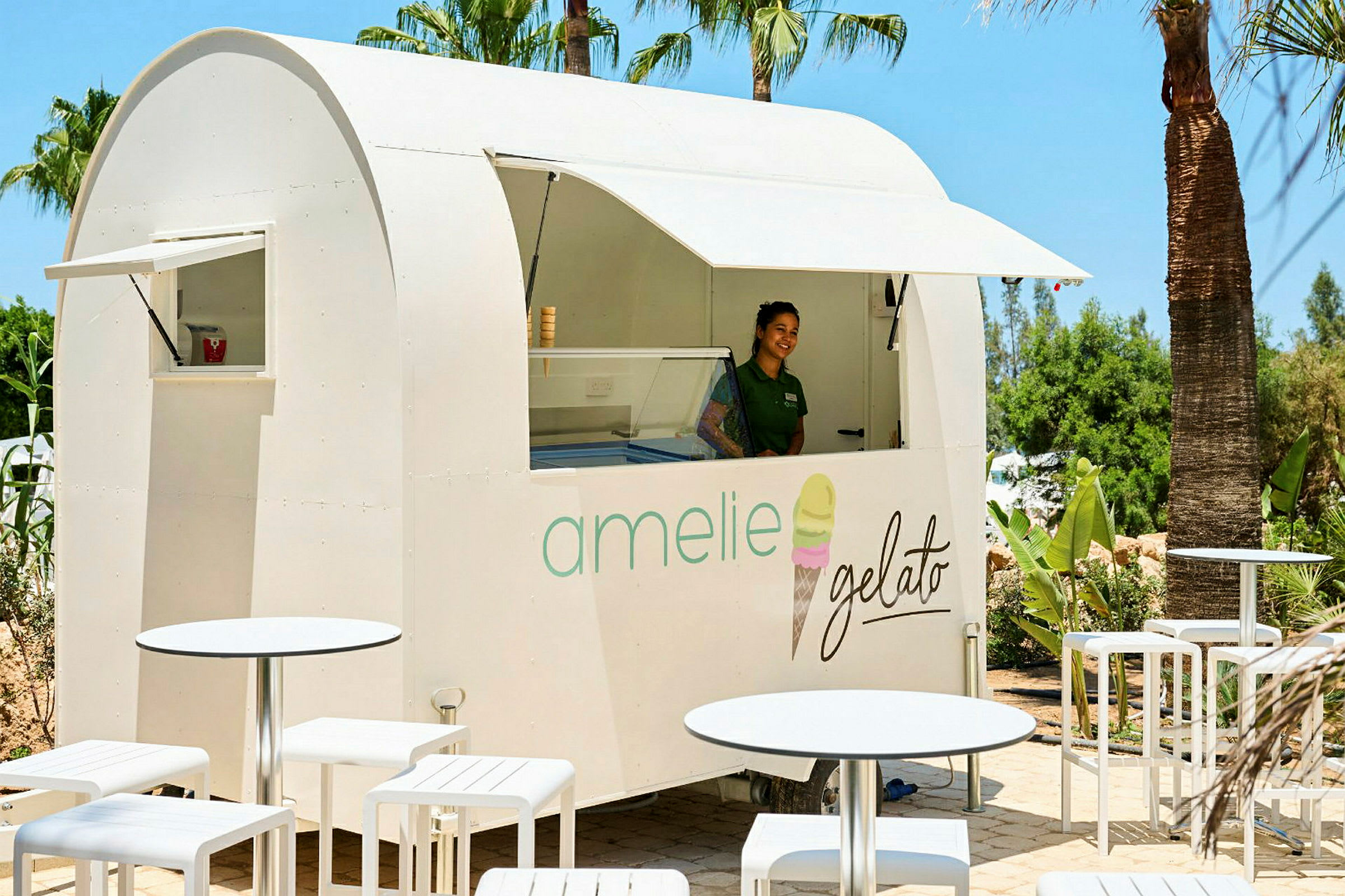 En hvit iskremkiosk med teksten "amelie gelato" står blant palmer, omgitt av hvite utemøbler. (AI-generert tekst)