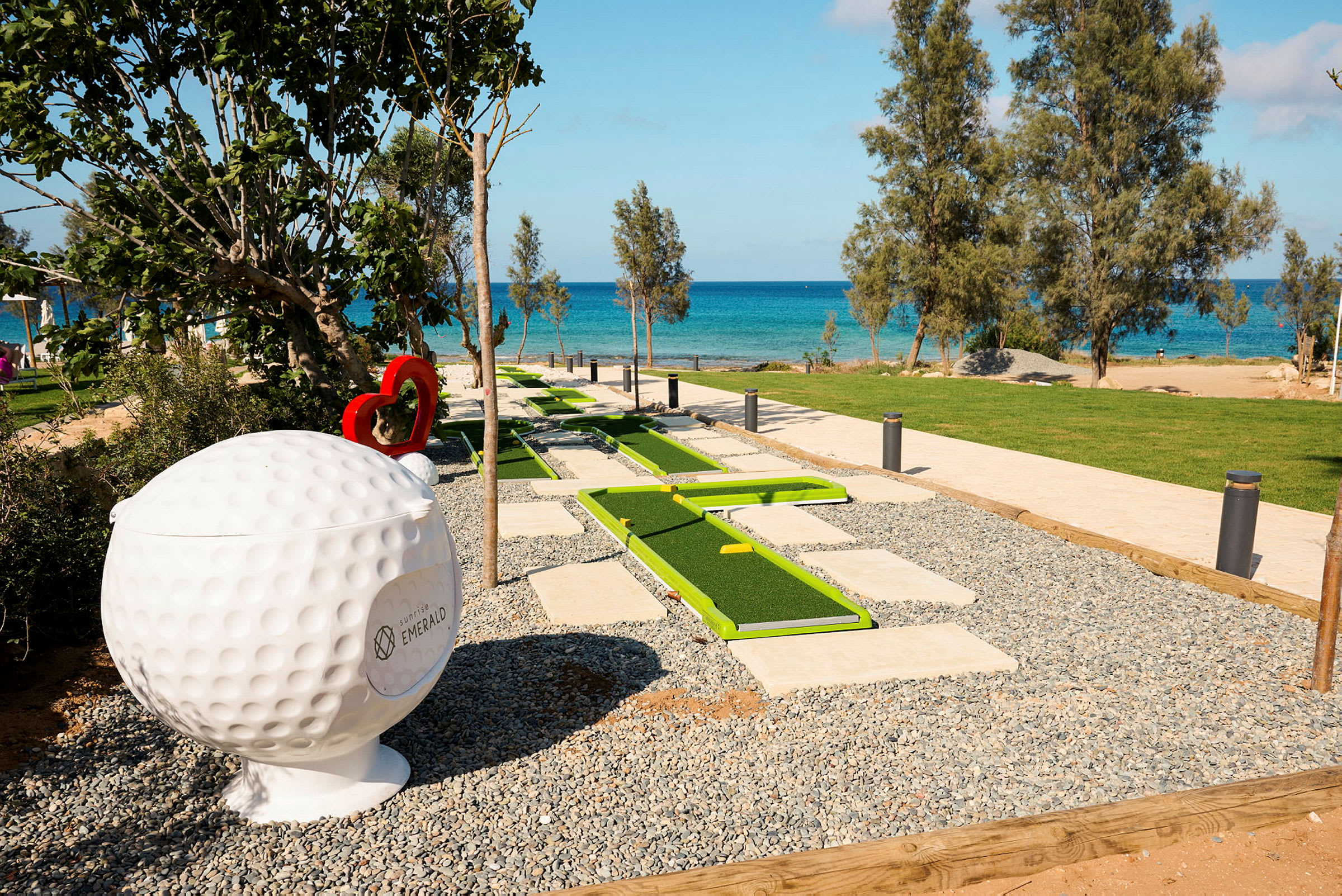 En minigolfbane med grønne baner ligger nær stranden med blått hav i bakgrunnen. En stor golfhullskulptur kan sees ved siden av. (AI-generert tekst)