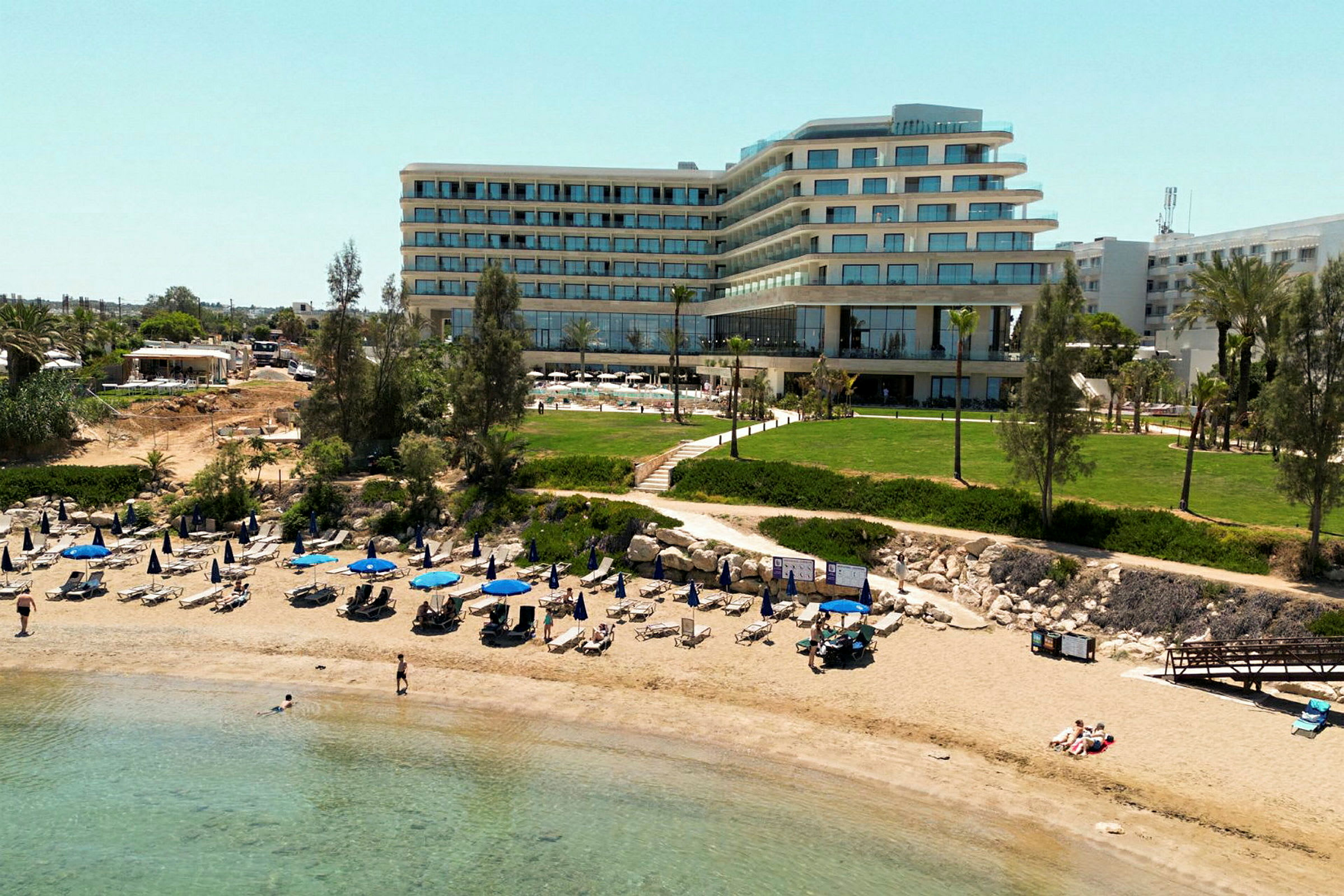 Bilde av en sandstrand med oppstilte parasoller og solsenger foran en moderne hotellbygning og grøntområder. (AI-generert tekst)