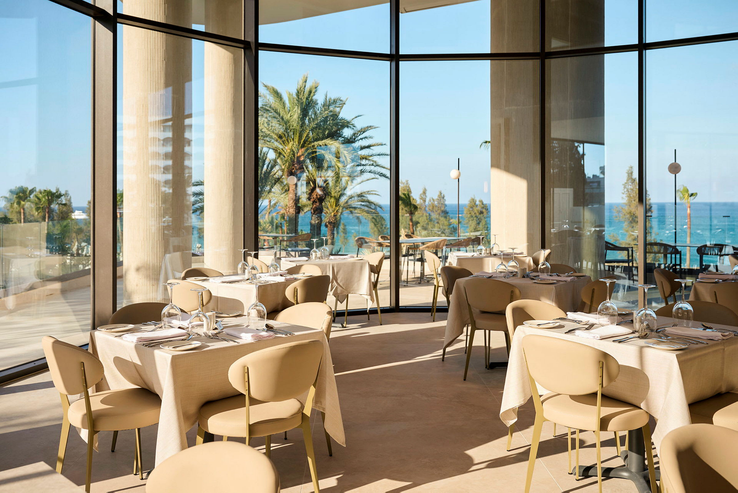 En lys og elegant restaurant med panoramavinduer som viser utsikt over palmer og havet. (AI-generert tekst)