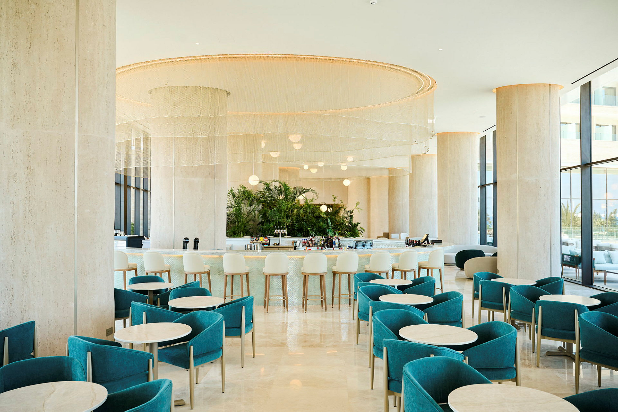En moderne hotellbar med turkise stoler og runde bord, en elegant bardisk i bakgrunnen og store vinduer som slipper inn dagslys. (AI-generert tekst)