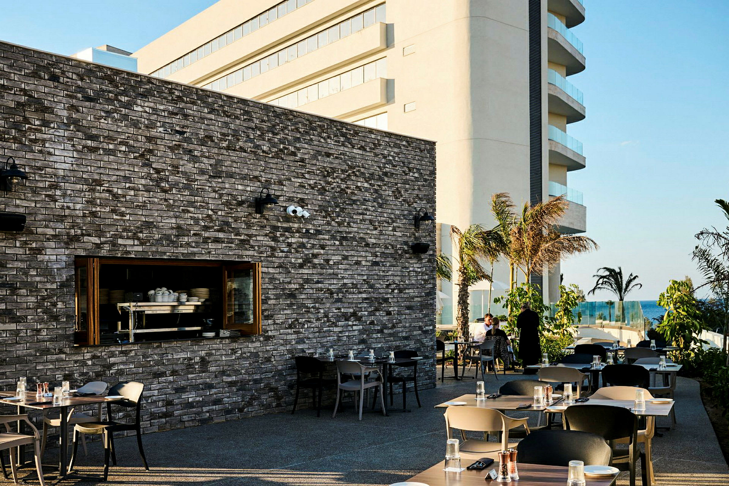 Uteområde ved en restaurant med bord og stoler. En stor bygning vises i bakgrunnen, med palmer og havet i bakgrunnen. (AI-generert tekst)