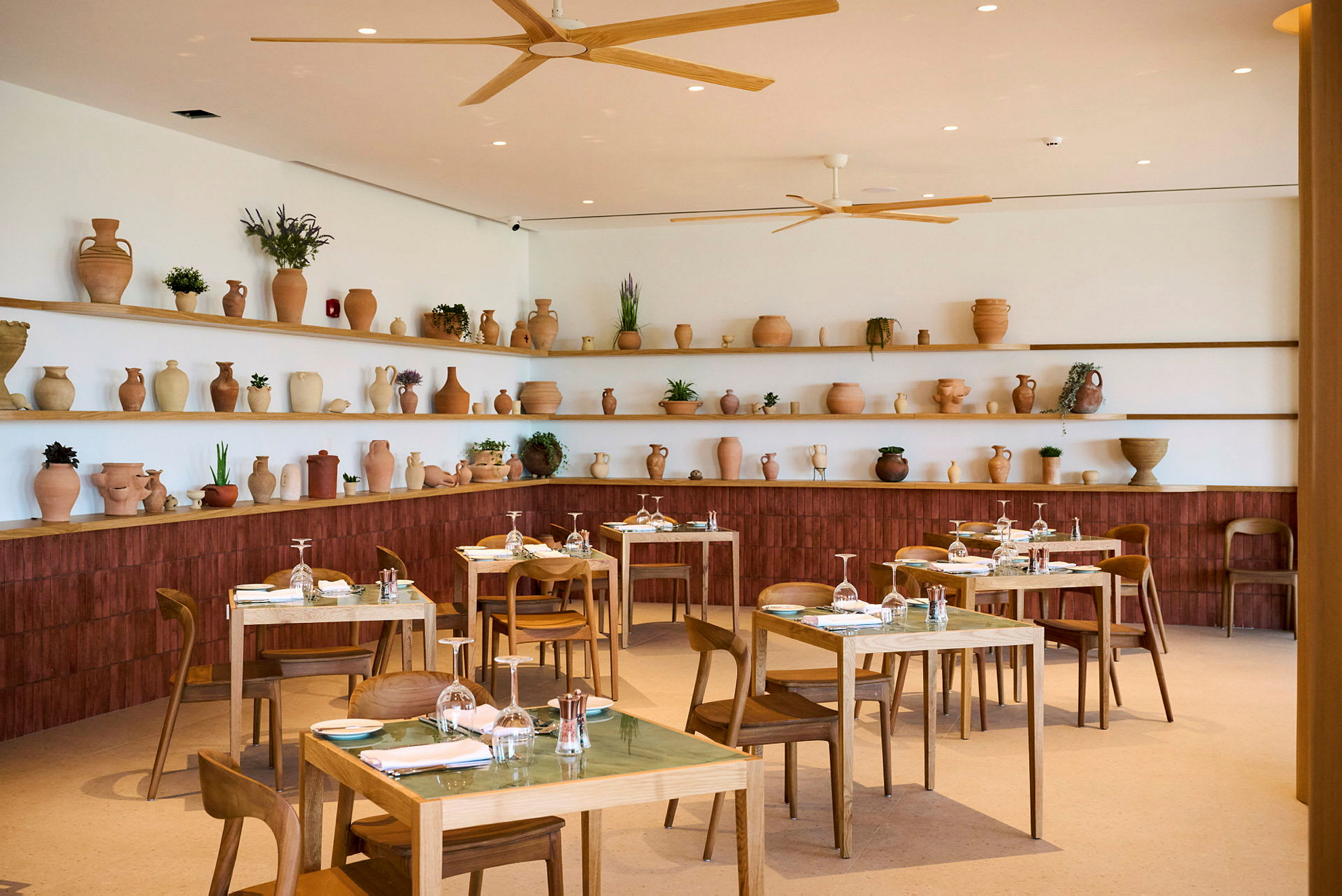 Bilde av et restaurantinteriør med trebord og stoler. Hyller på veggen bak er dekorert med terrakottakrukker og planter. (AI-generert tekst)