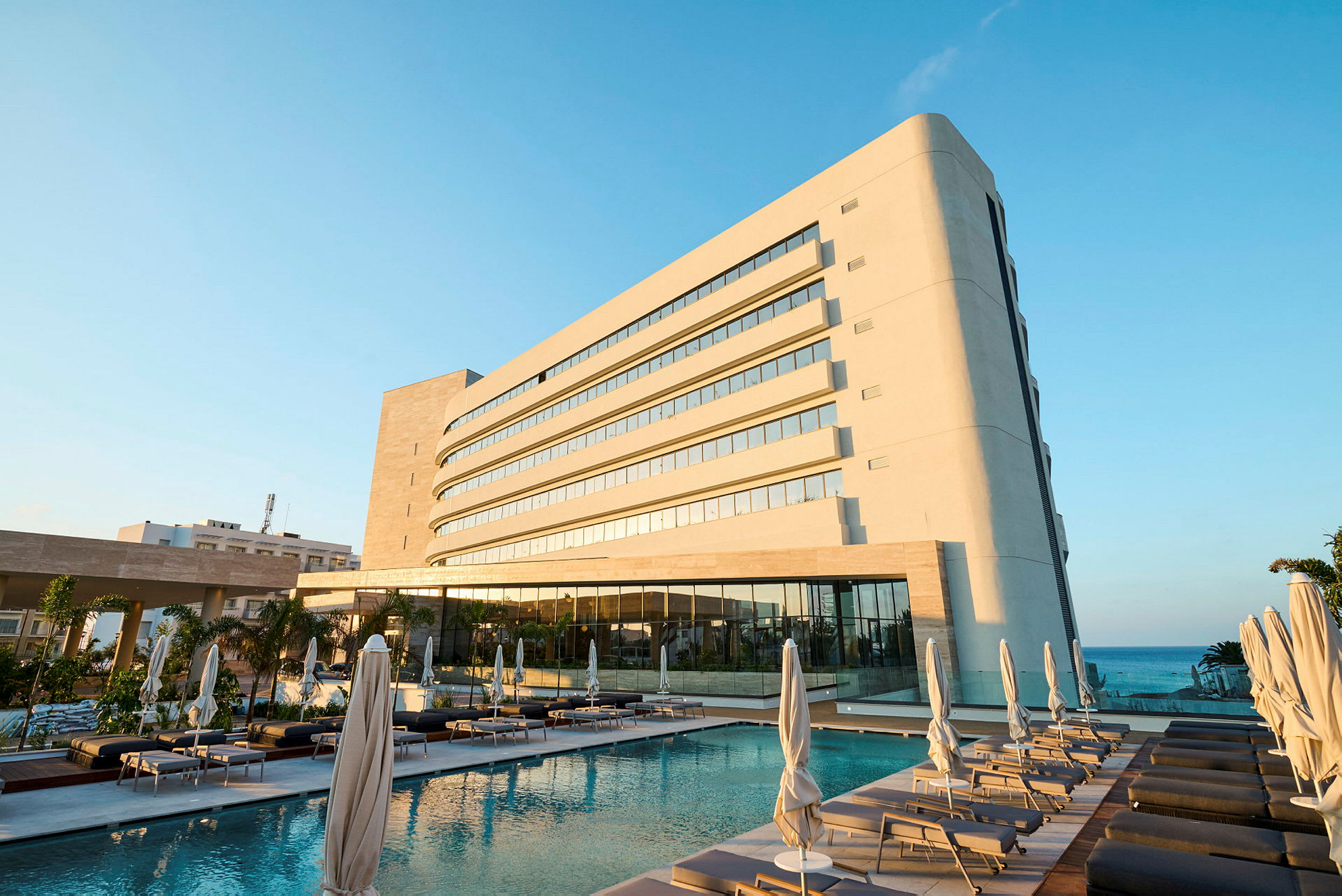En moderne hotellbygning ved siden av et basseng med solsenger og parasoller. Havet kan sees i bakgrunnen. (AI-generert tekst)
