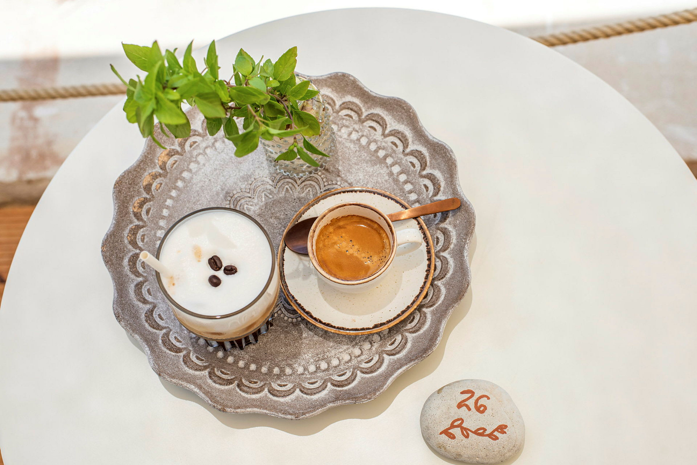 Et detaljbilde av et brett med en kopp kaffe, en milkshake med kaffe og en liten glassvase med grønne blader på et bord. (AI-generert tekst)