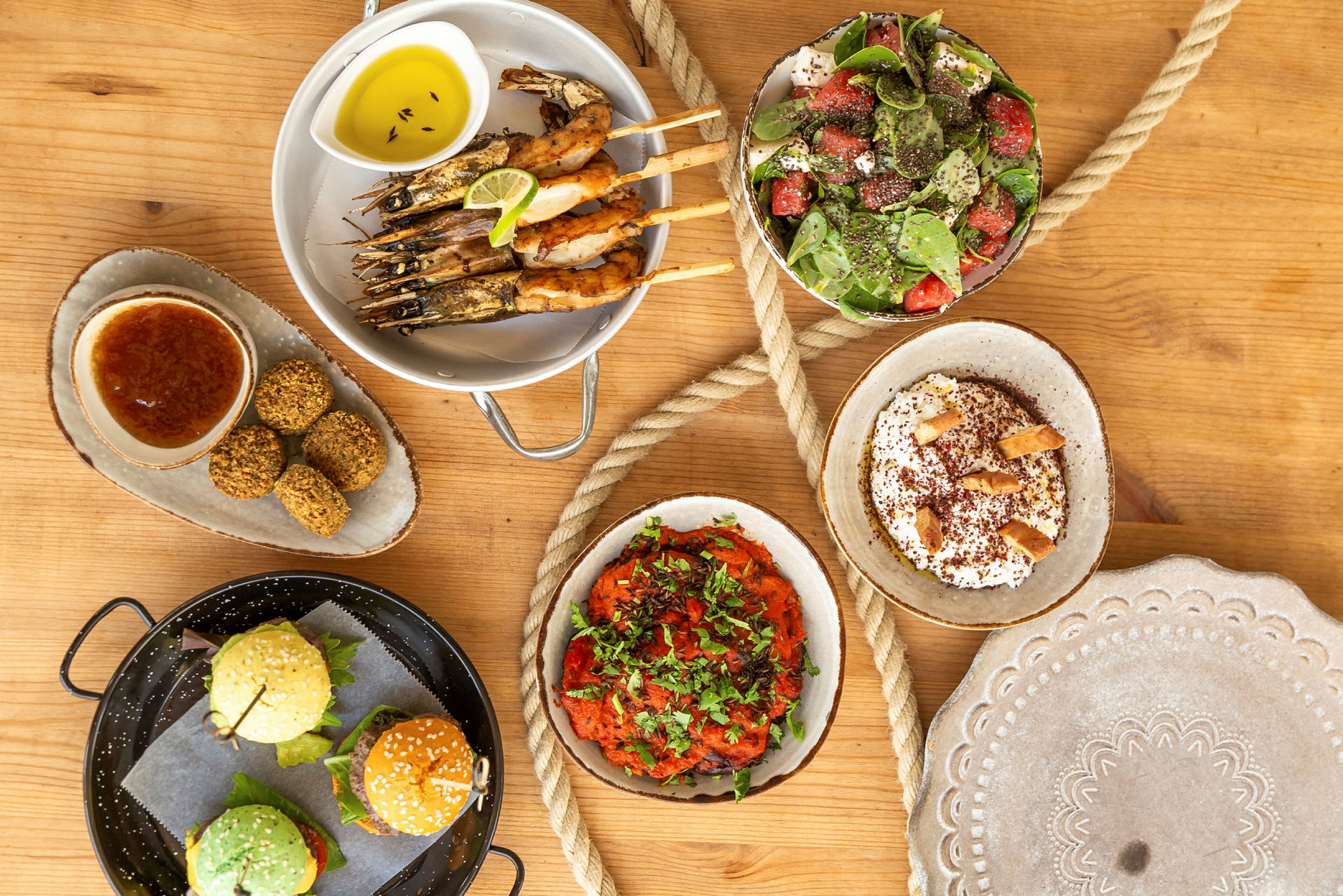 En rekke ulike matretter som reker på spyd, salat, falafel, burgere, samt dippsauser og tilbehør på et trebord. (AI-generert tekst)