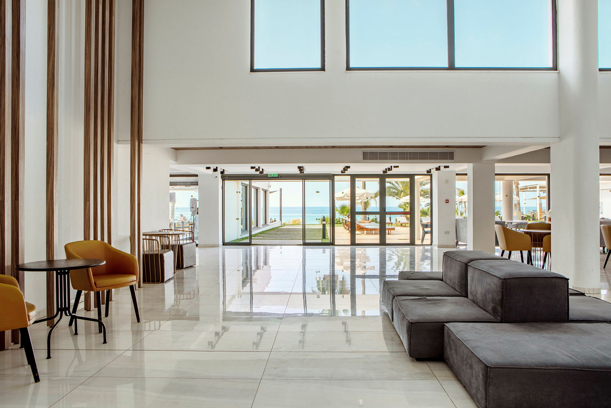 En moderne hotellobby med lys innredning, sitteområder og glassdører som fører til et utendørs basseng og havet. (AI-generert tekst)